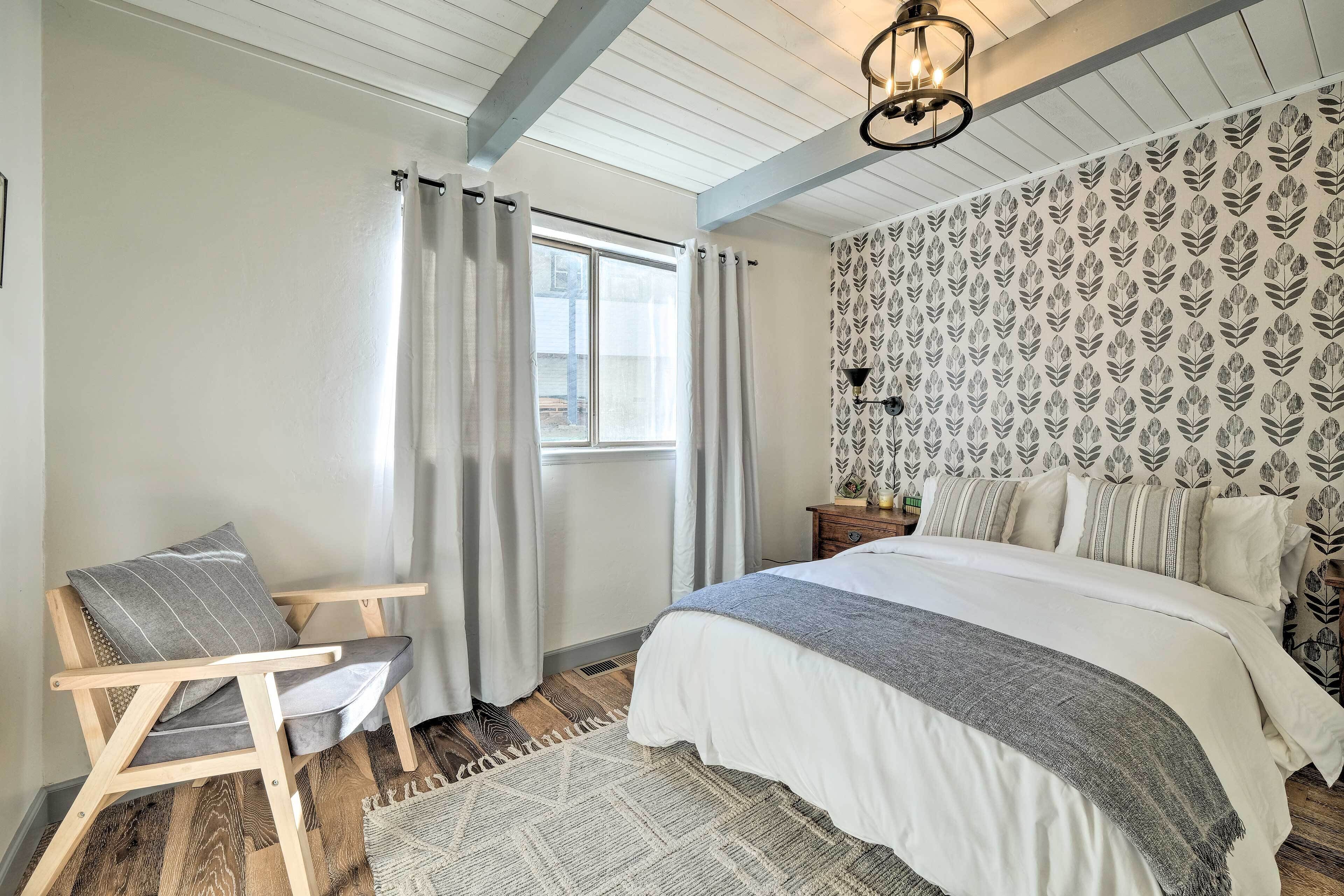 A bedroom with a bed, a chair, a window with curtains, a patterned wall, and a ceiling light.