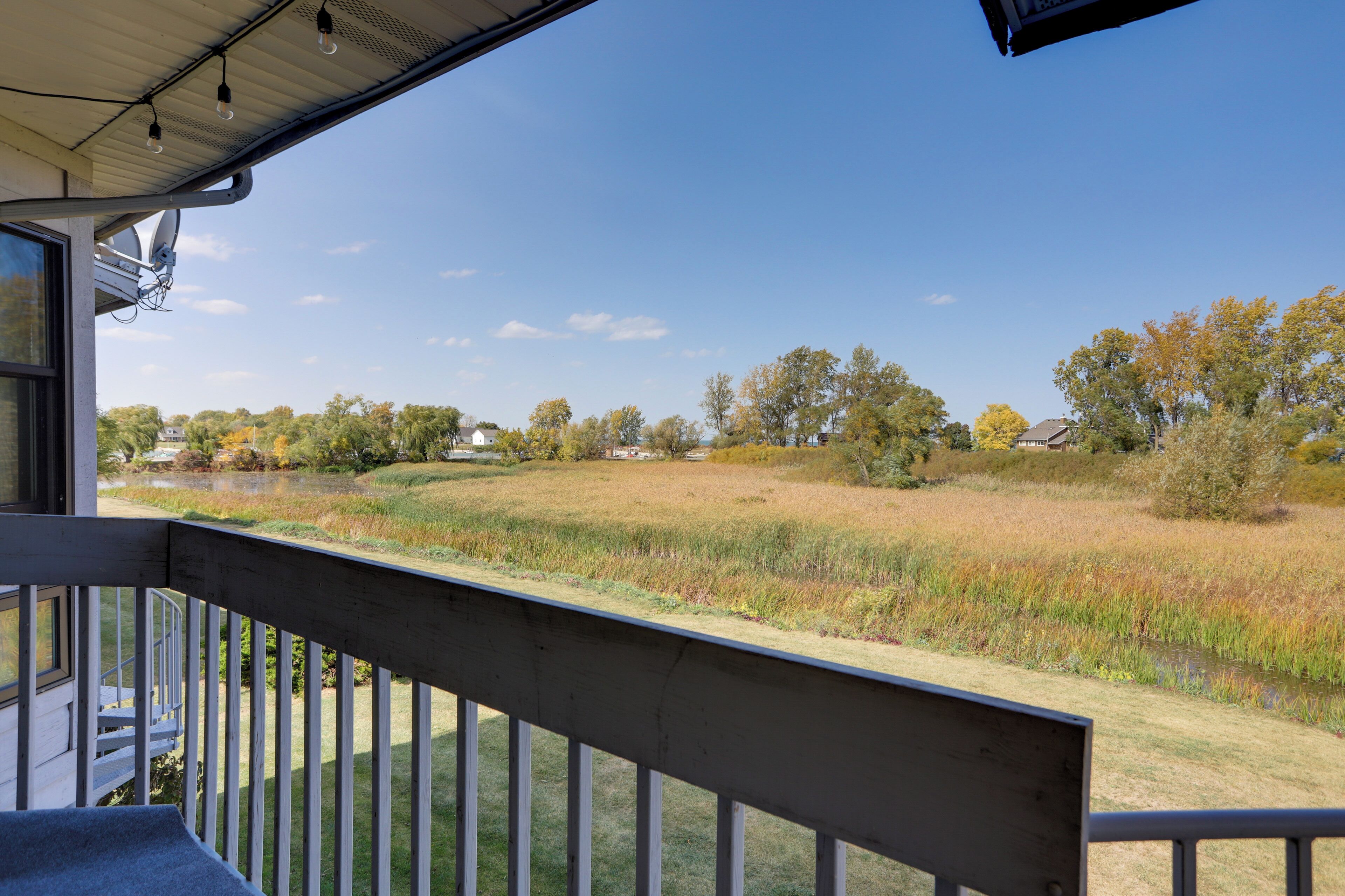 A view from a balcony overlooking a grassy field with a pond, trees, and houses in the distance.
