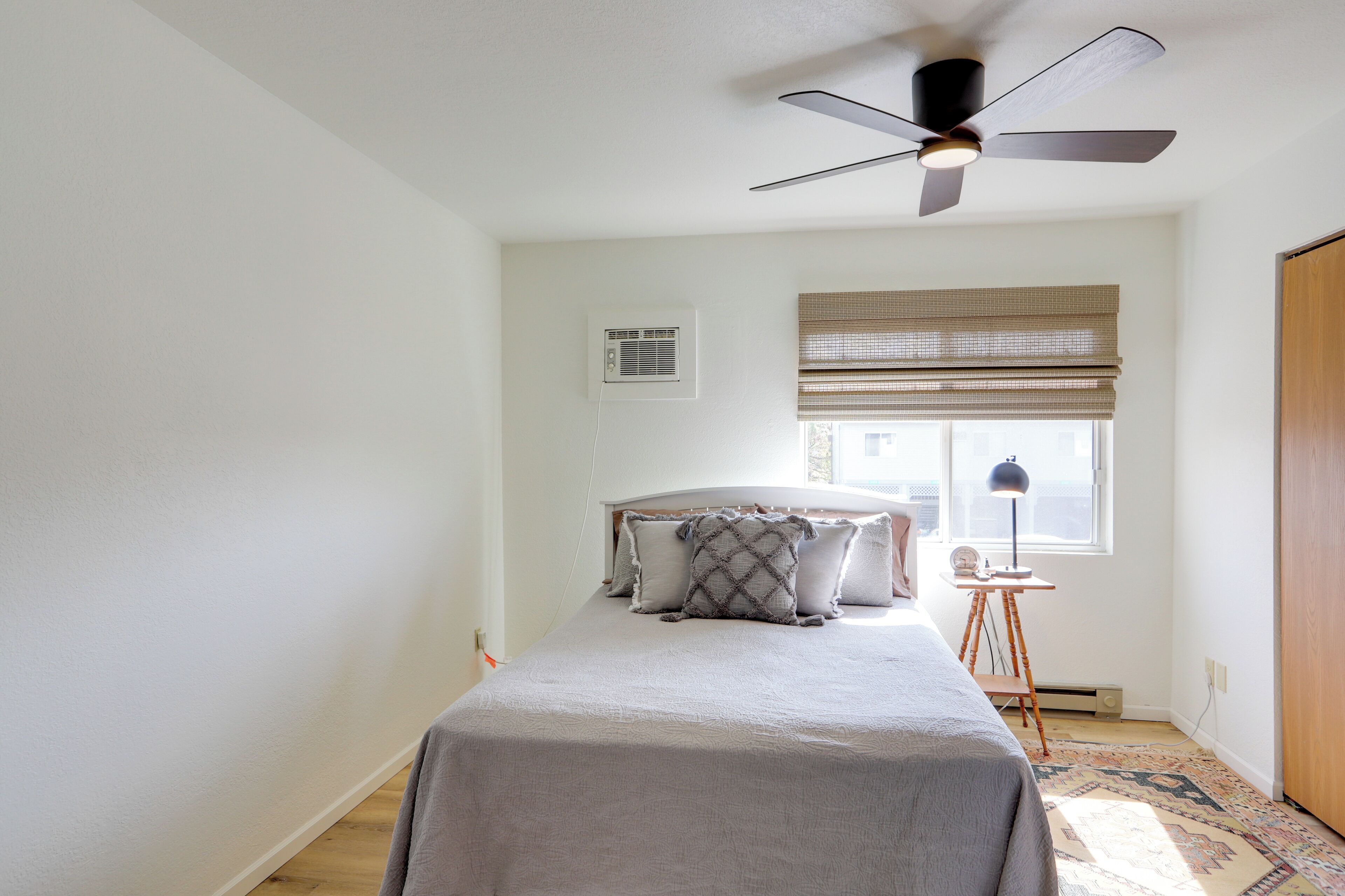 Ein Schlafzimmer mit Bett, Deckenventilator, Fenster mit Jalousien, Schreibtisch mit Lampe und Heizkörper.