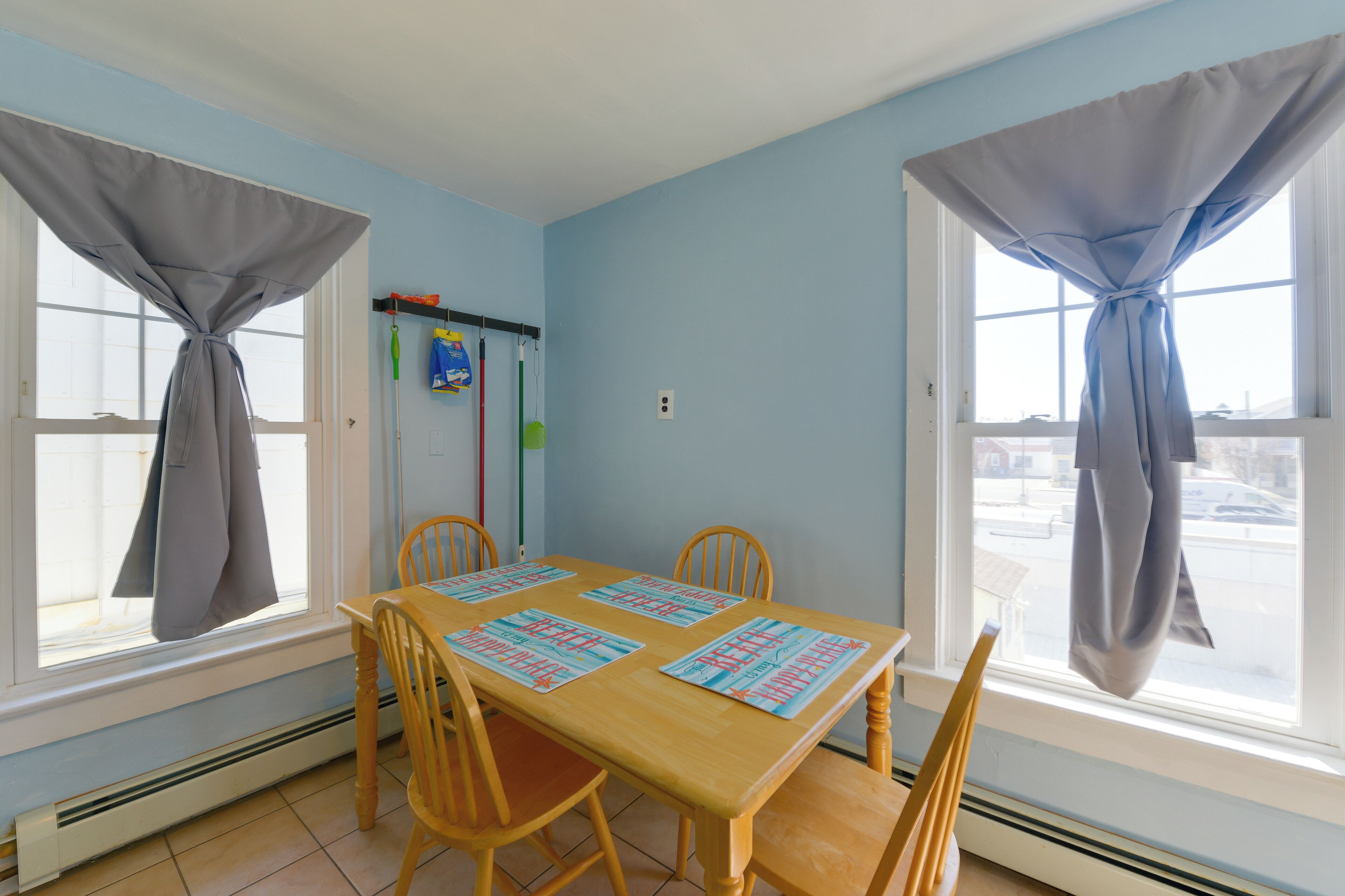 Comedor con mesa de madera y cuatro sillas, dos ventanas con cortinas y un estante en la pared con artículos de limpieza.