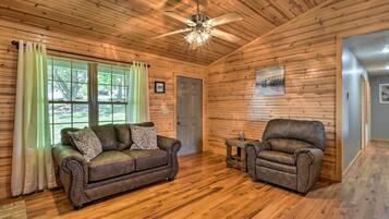 A cozy room with wooden walls and floors, a ceiling fan, a brown leather sofa, a smaller leather armchair, and a small table with a vase.