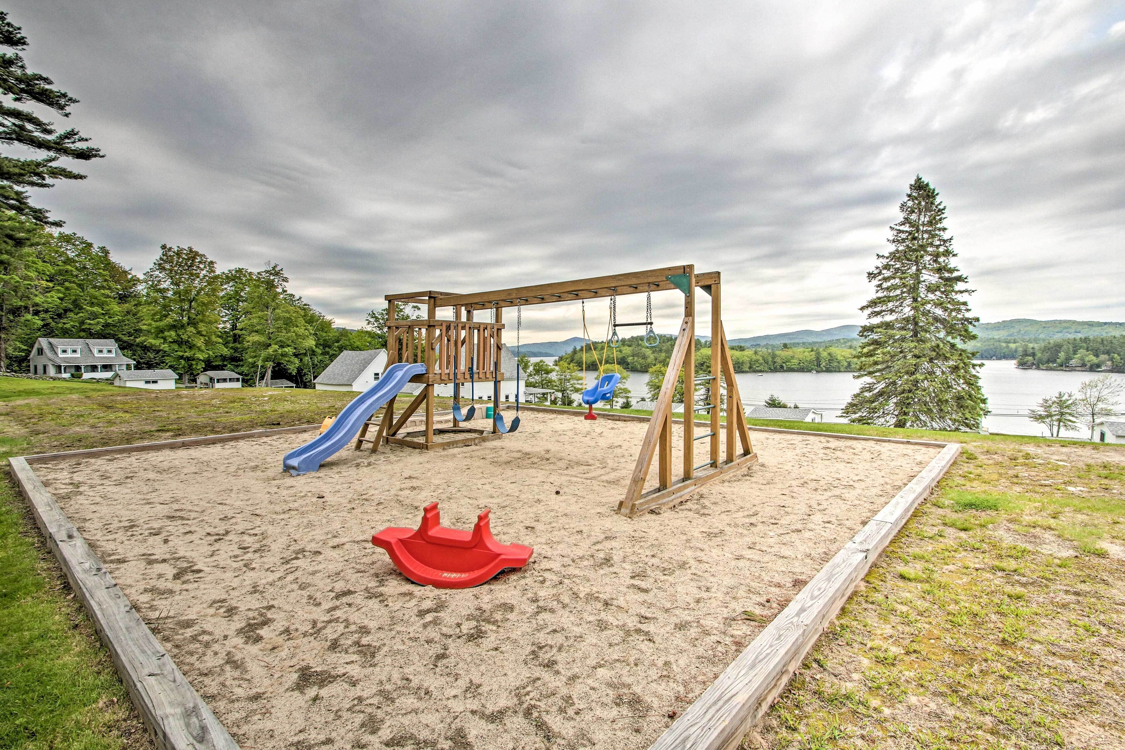 Ein Spielplatz mit Schaukeln und Rutschen, umgeben von Sand und einer Holzumrandung.
