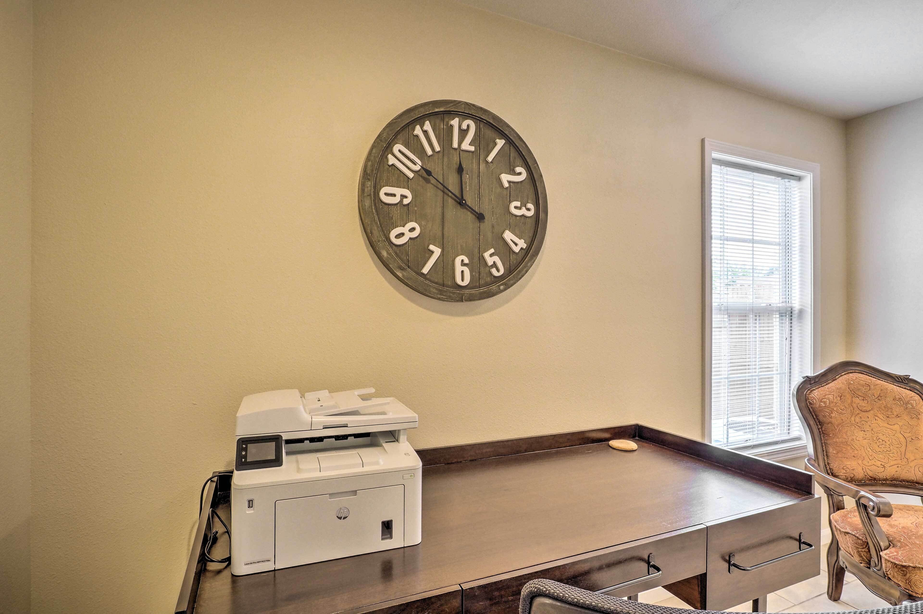 A room with a wooden desk, a printer, a clock, and a window with blinds.