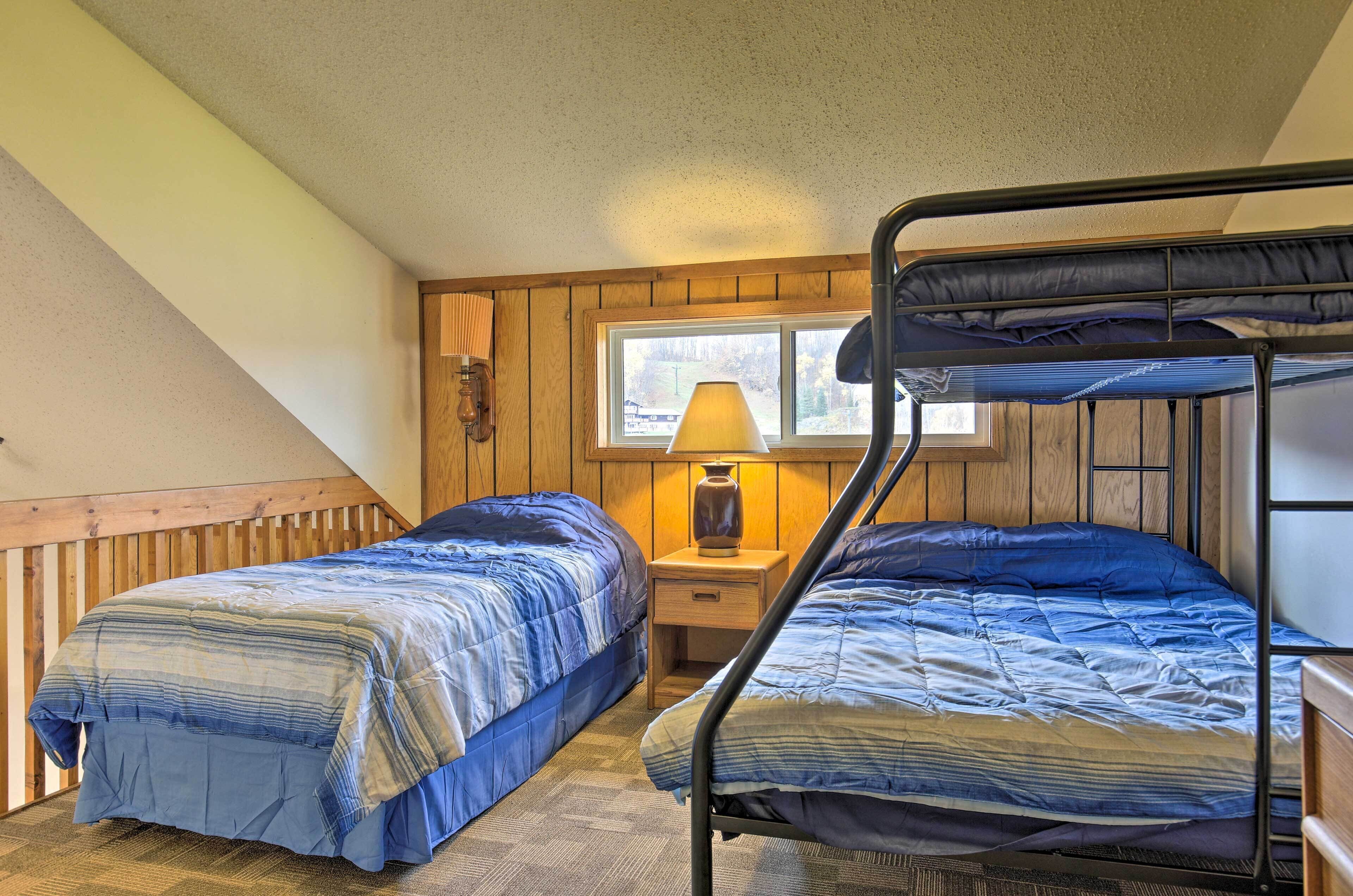 Une chambre avec deux lits superposés, une tête de lit en bois, une table de chevet avec une lampe et une fenêtre donnant sur des arbres.