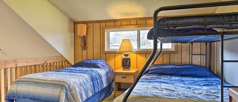 Une chambre avec deux lits superposés, une tête de lit en bois, une table de chevet avec une lampe et une fenêtre donnant sur des arbres.