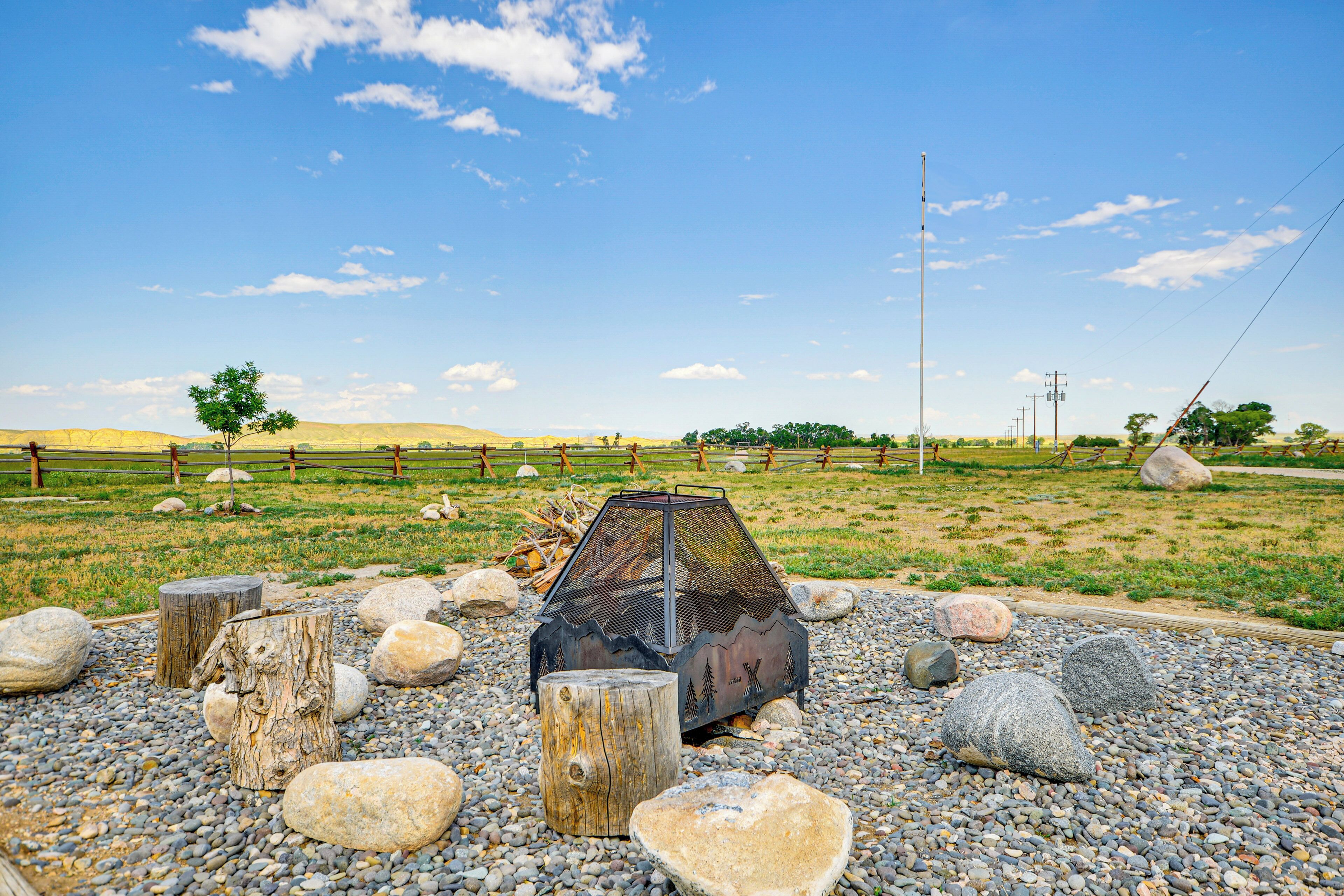 A metal fire pit surrounded by rocks and wooden stumps in a rural outdoor setting.