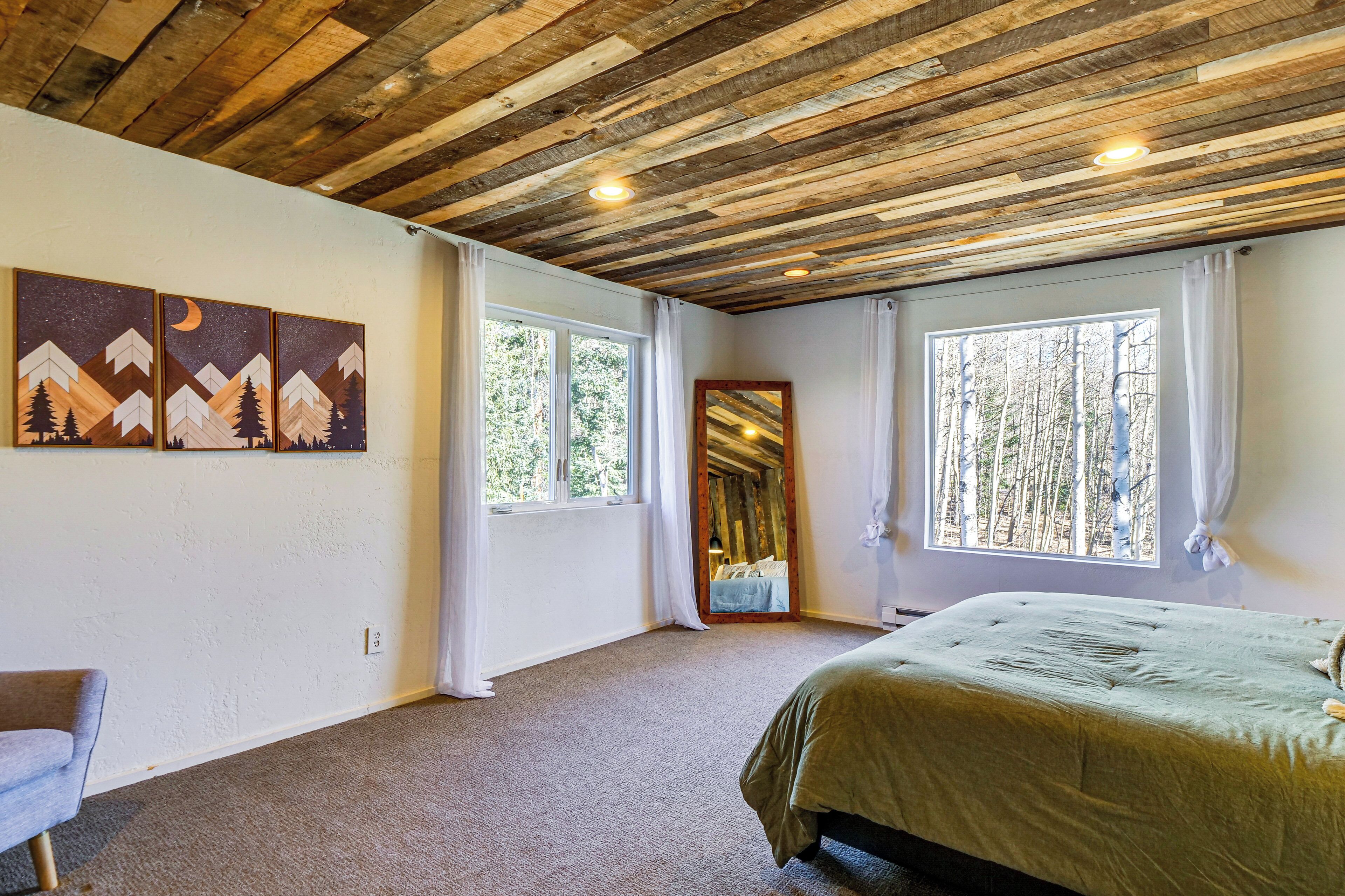 Un dormitorio con techo de madera, una cama, dos ventanas con cortinas y un cuadro en la pared.