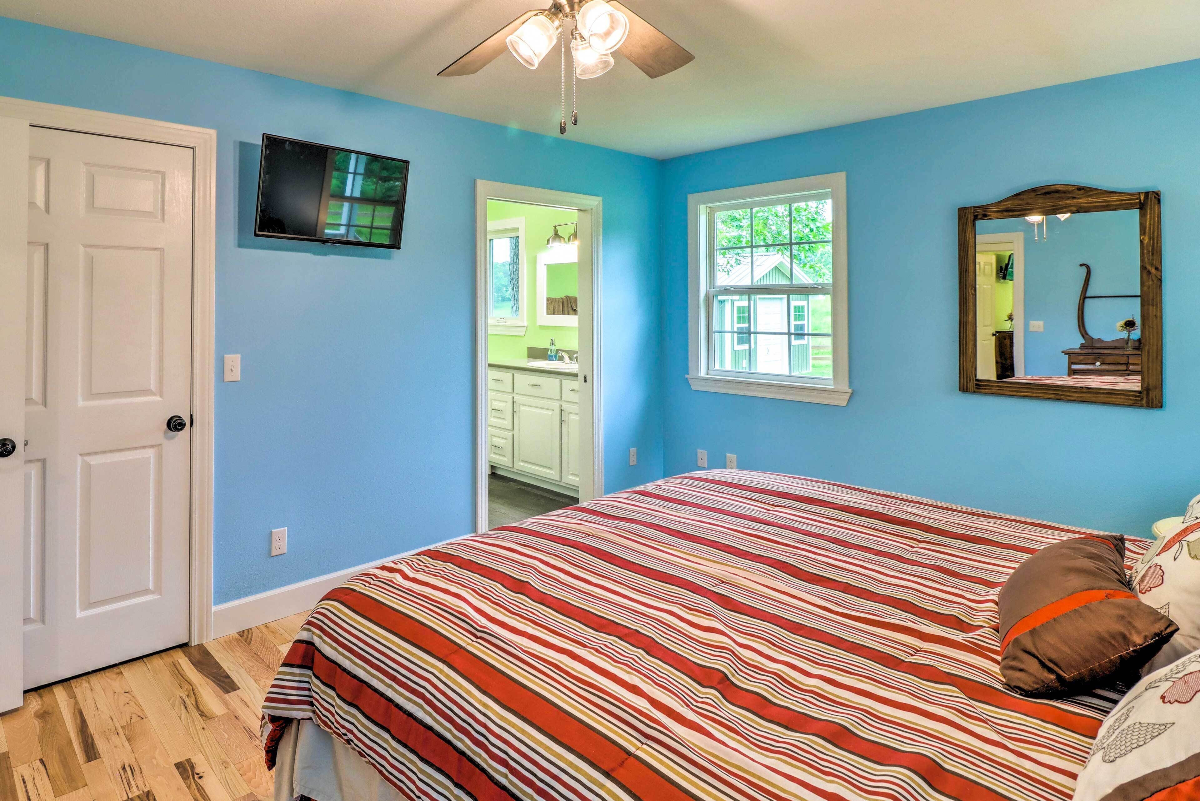 A bedroom with a bed, a ceiling fan, a TV, a window, and a door.