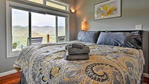 A bedroom with a large bed, a view of the outdoors, a wall-mounted lamp, and a framed picture. - Private Blue Ridge Home w/ Hot Tub, Mountain Views (Marshall)