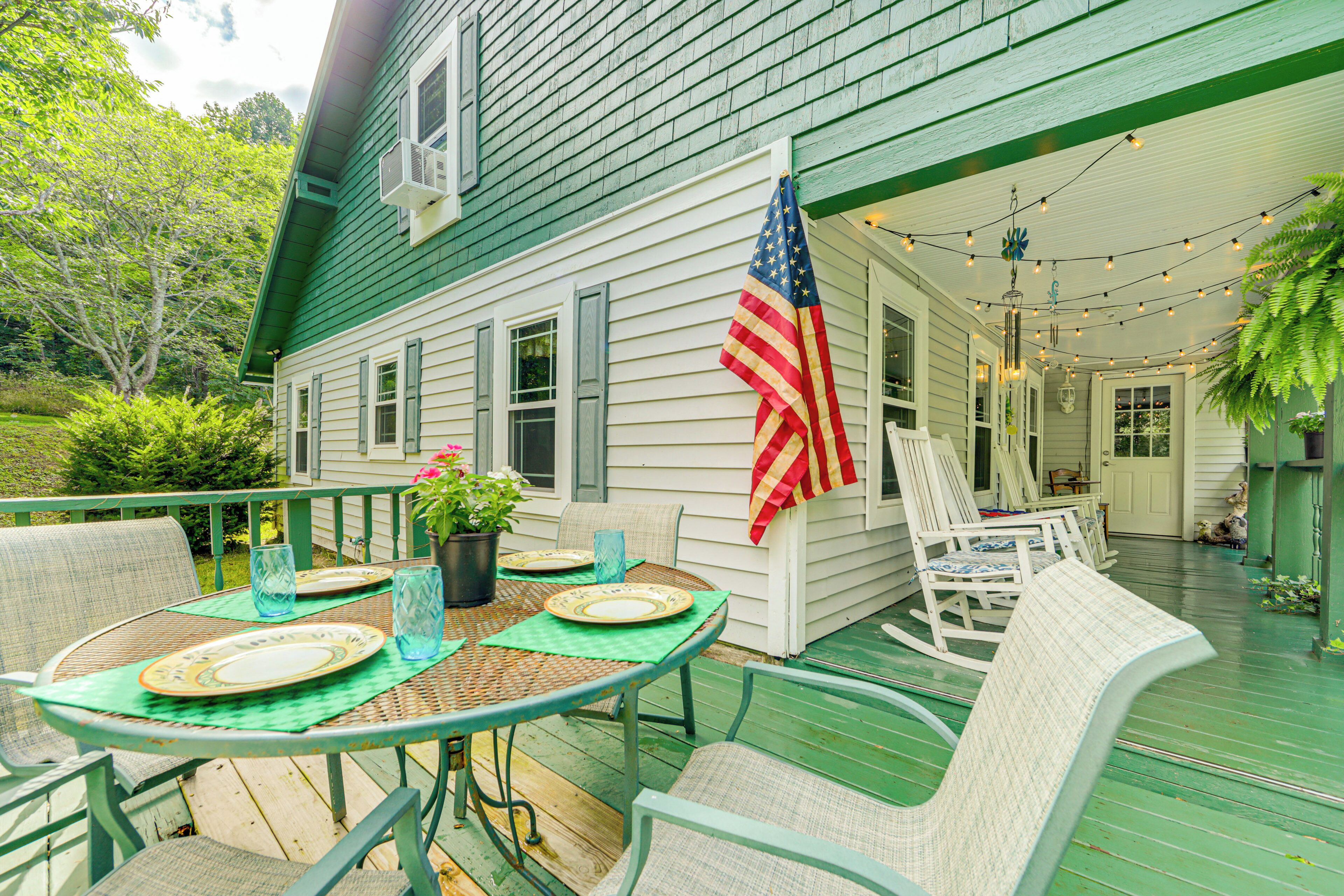 Une terrasse en bois peinte en vert, avec une table dressée pour quatre personnes, entourée de chaises en osier, et un drapeau américain accroché au mur.