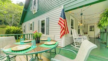 Une terrasse en bois peinte en vert, avec une table dressée pour quatre personnes, entourée de chaises en osier, et un drapeau américain accroché au mur.