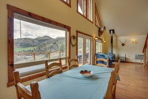 Un comedor con una mesa dispuesta para dos personas, sillas de madera y vistas a un paisaje montañoso a través de amplios ventanales.