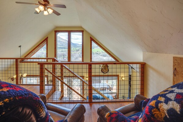 A cozy loft bedroom with a high ceiling, a wooden staircase, and large windows offering a view of the outdoors.