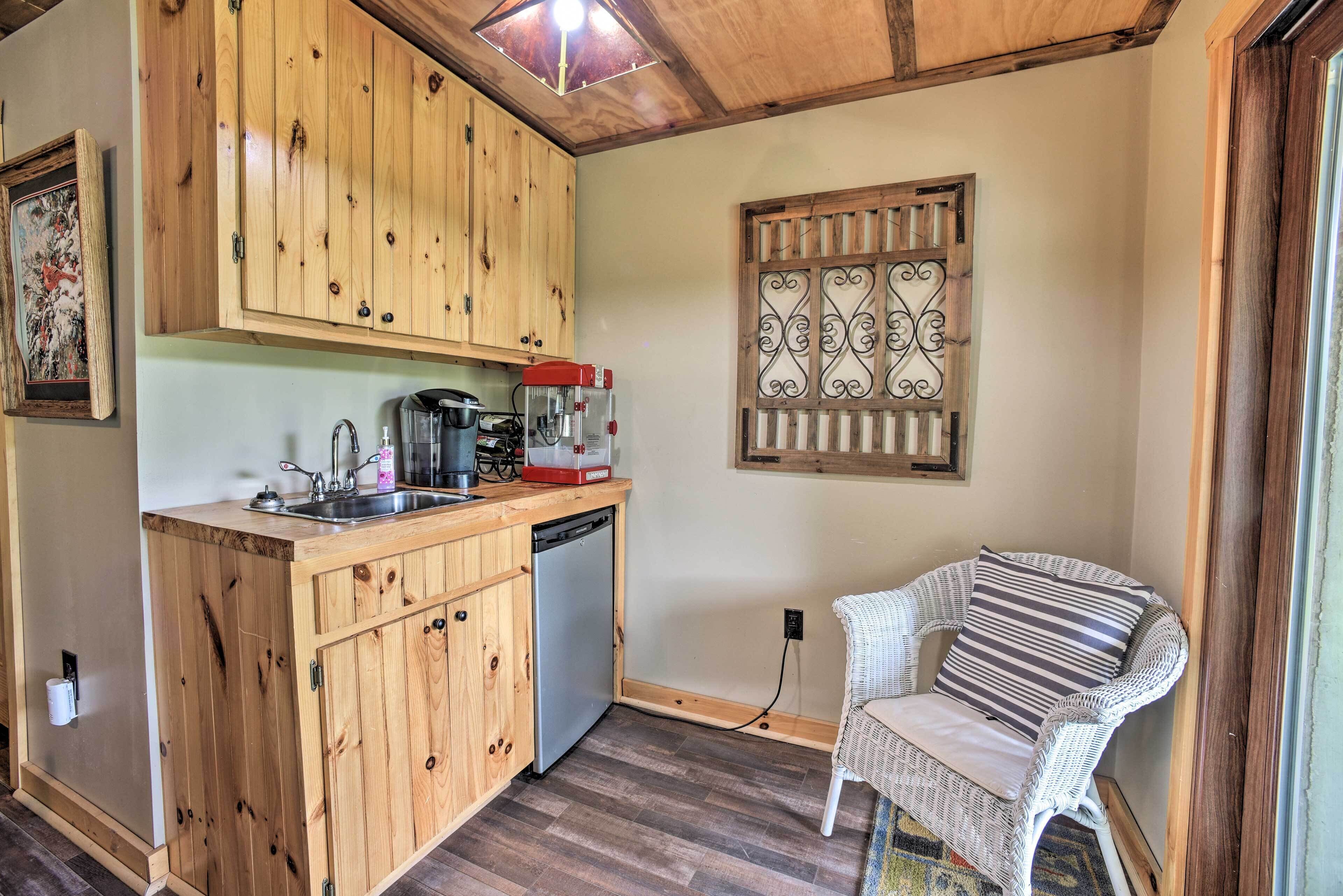 A compact kitchen with wooden cabinets, a coffee maker, and a dishwasher. There is a wicker chair and a decorative metal screen on the wall.