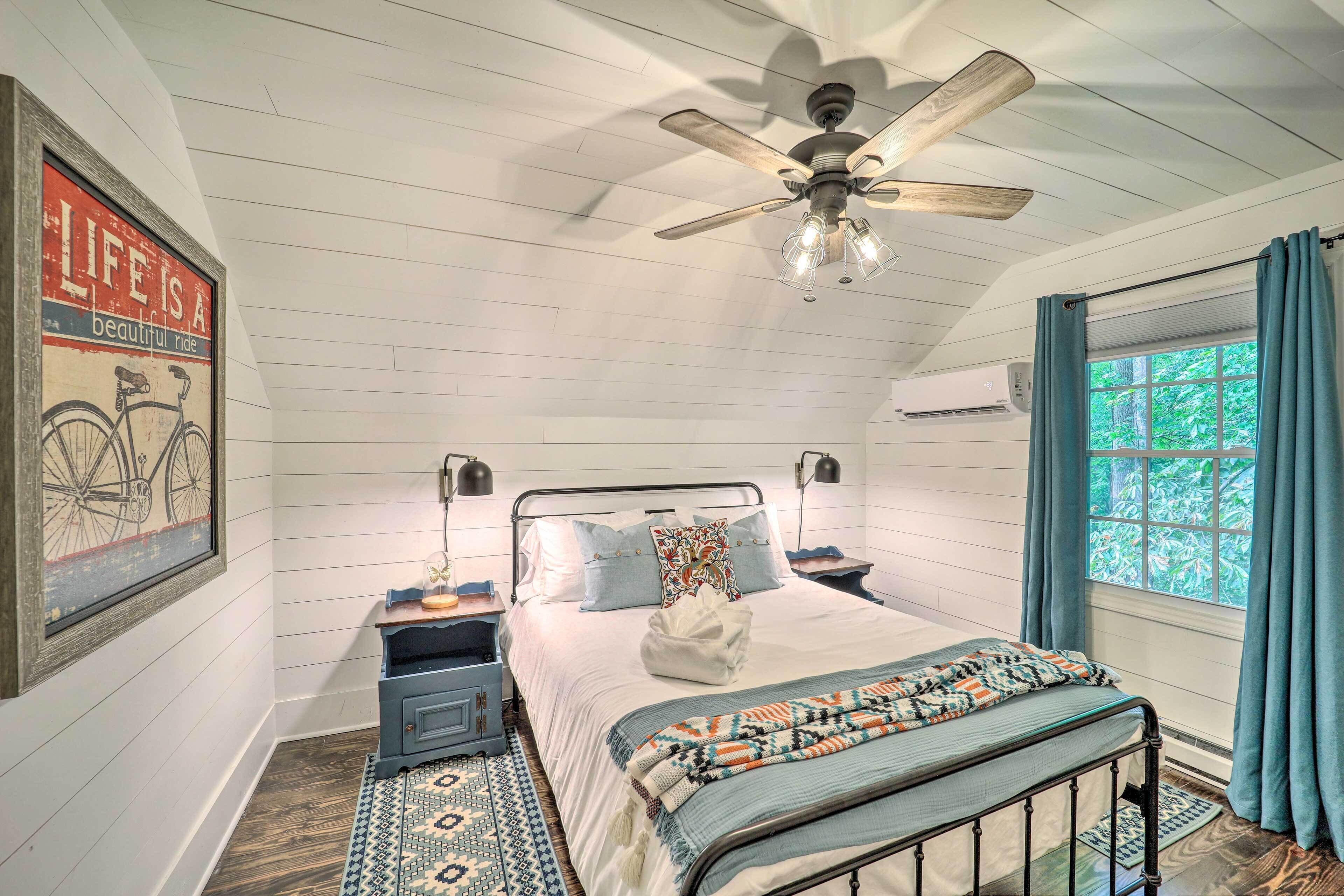 Ein Schlafzimmer mit einem Bett, einem Deckenventilator, einem Fenster mit Vorhängen, einer Wandklimaanlage und einem gerahmten Bild an der Wand.