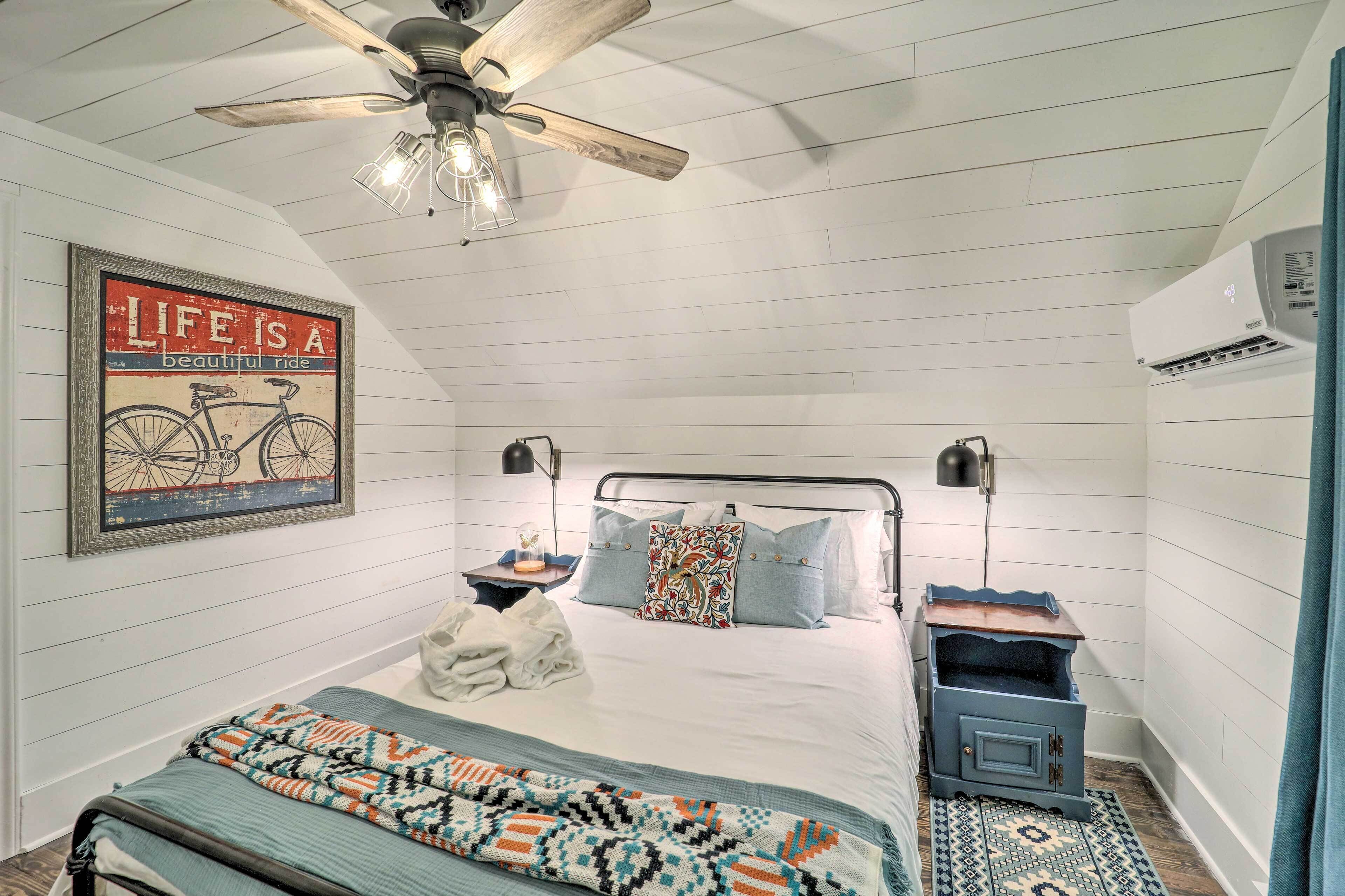 A bedroom with a bed, a ceiling fan, a wall-mounted air conditioner, and a framed inspirational quote.