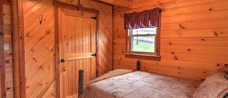 A wooden room with a bed, a window with a checkered valance, and a door.