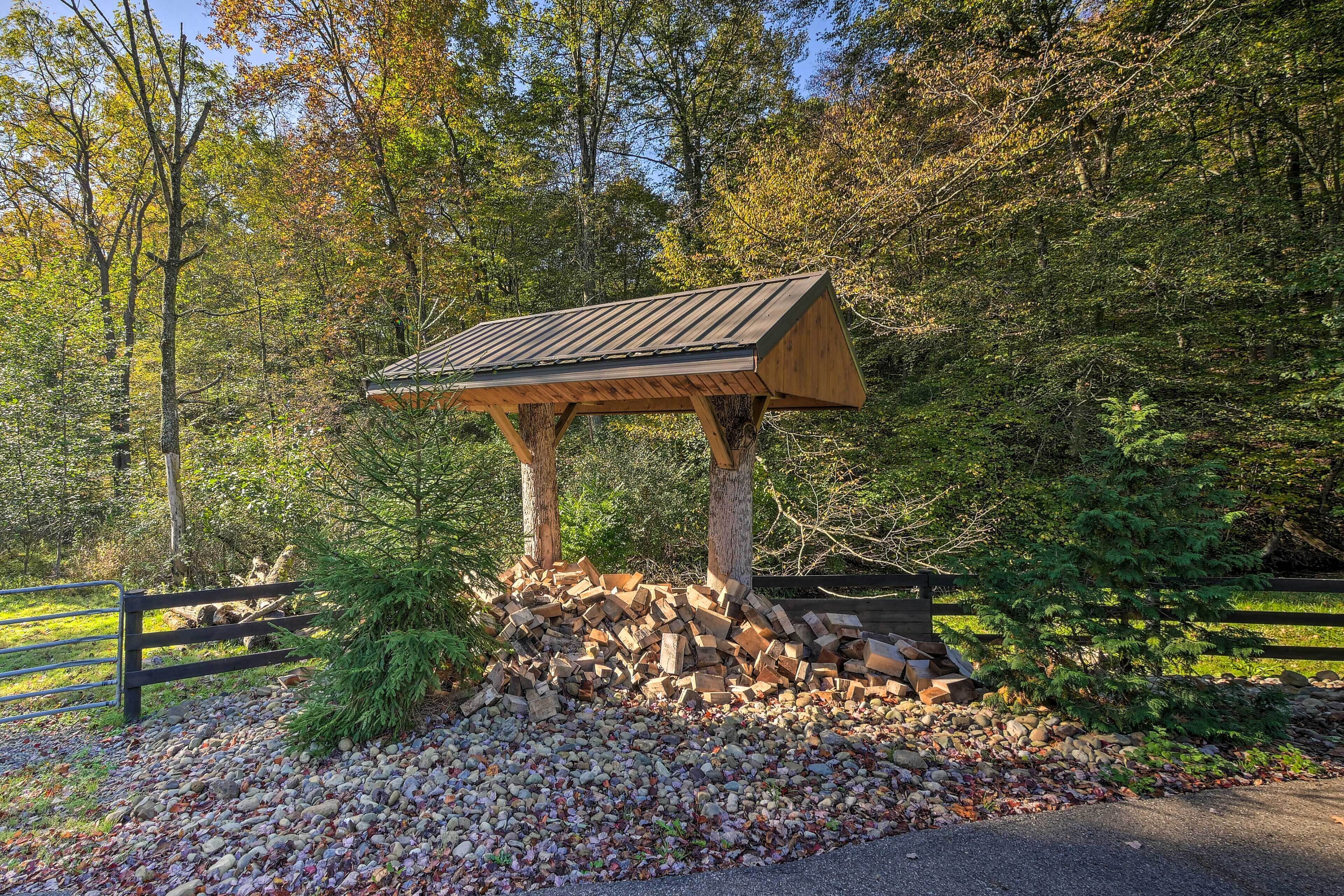 Un kiosque en bois doté d’un toit incliné, entouré d’une pile de bois de chauffage et de rochers, situé dans une zone boisée avec une clôture en arrière-plan.
