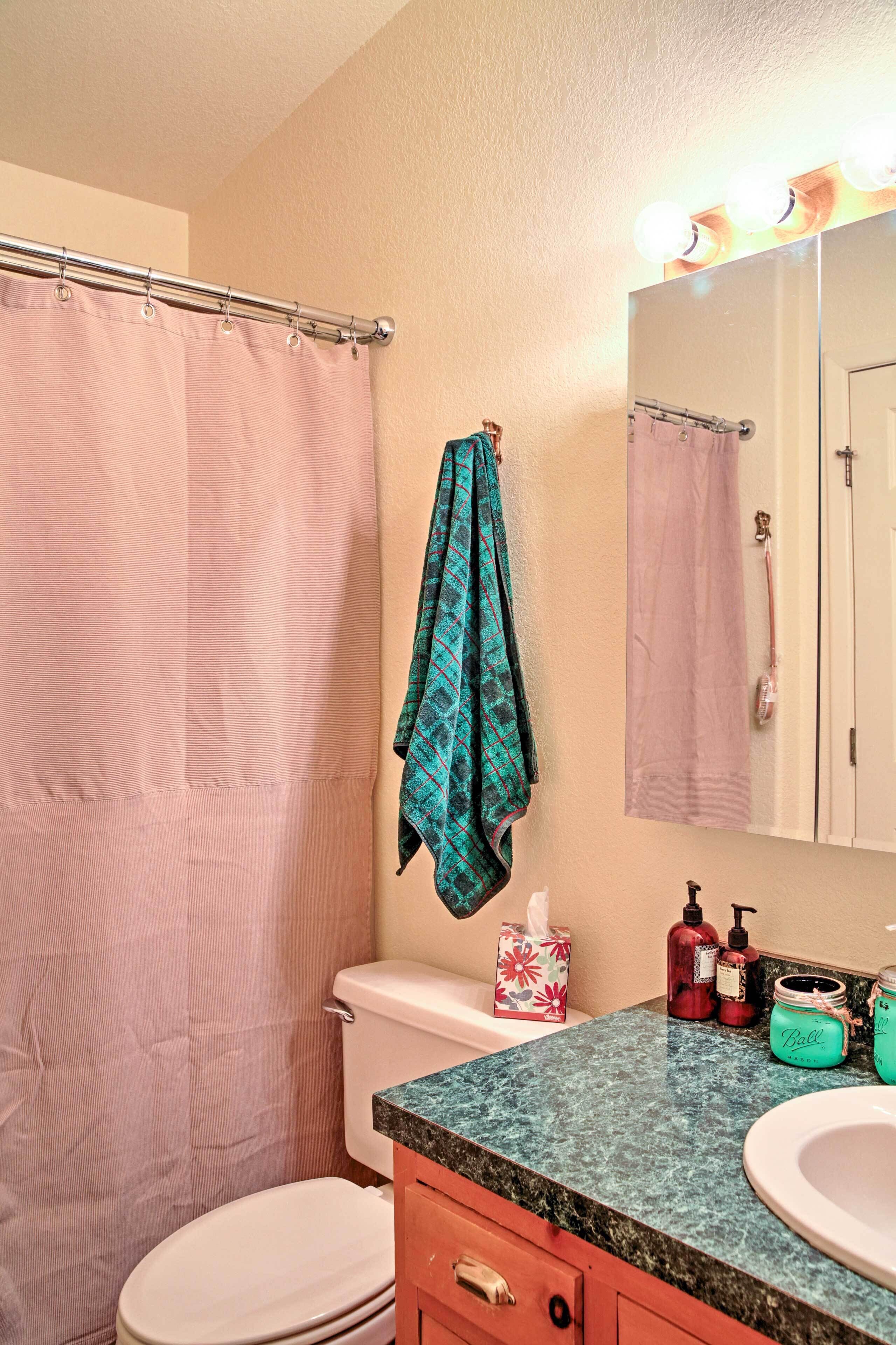 A bathroom with a pink shower curtain, a toilet, a sink with a green countertop, and a mirror with lights above it.
