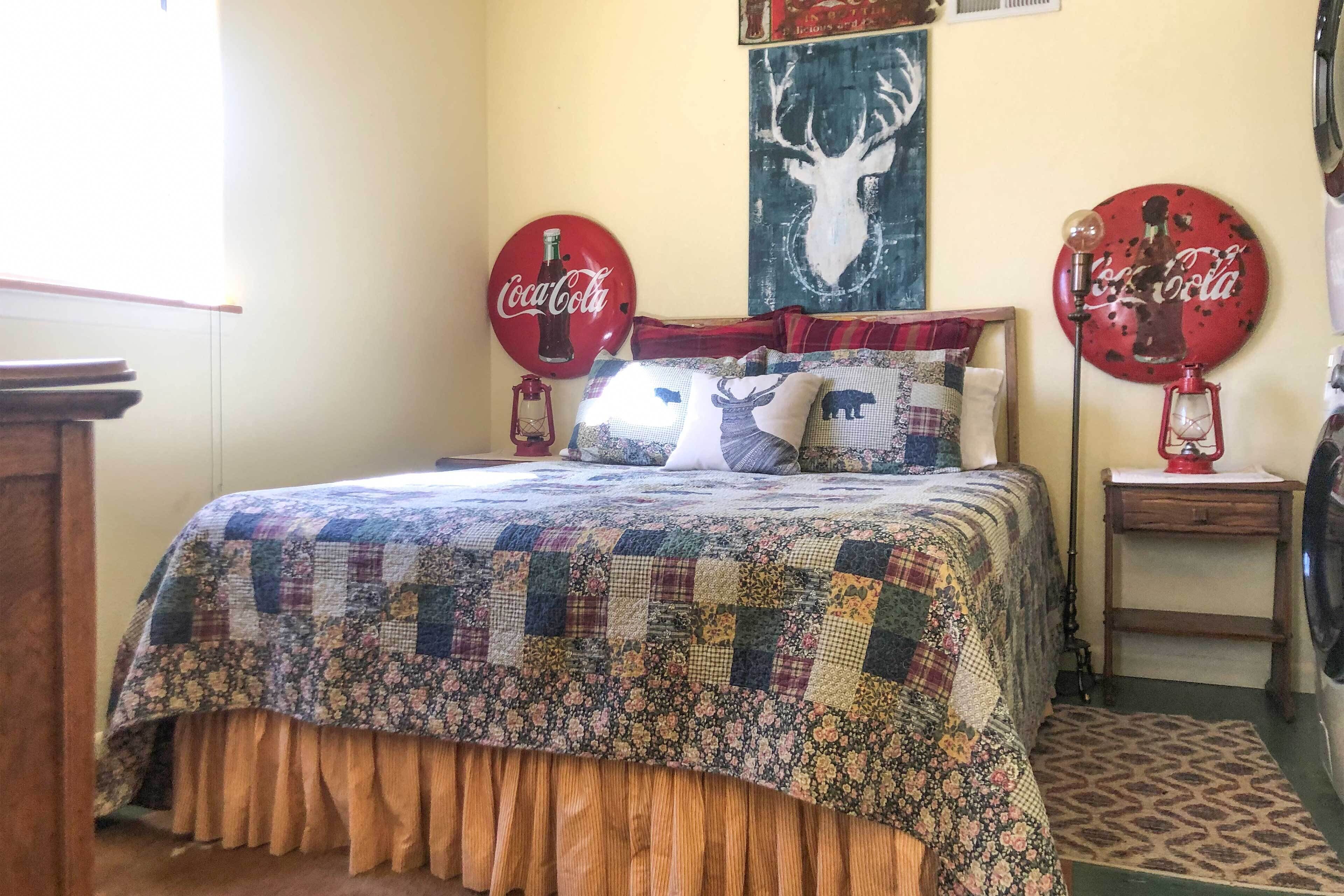 Un dormitorio con colcha acolchada, dos letreros de Coca-Cola, un cartel de un ciervo y una lámpara sobre una mesita de noche.