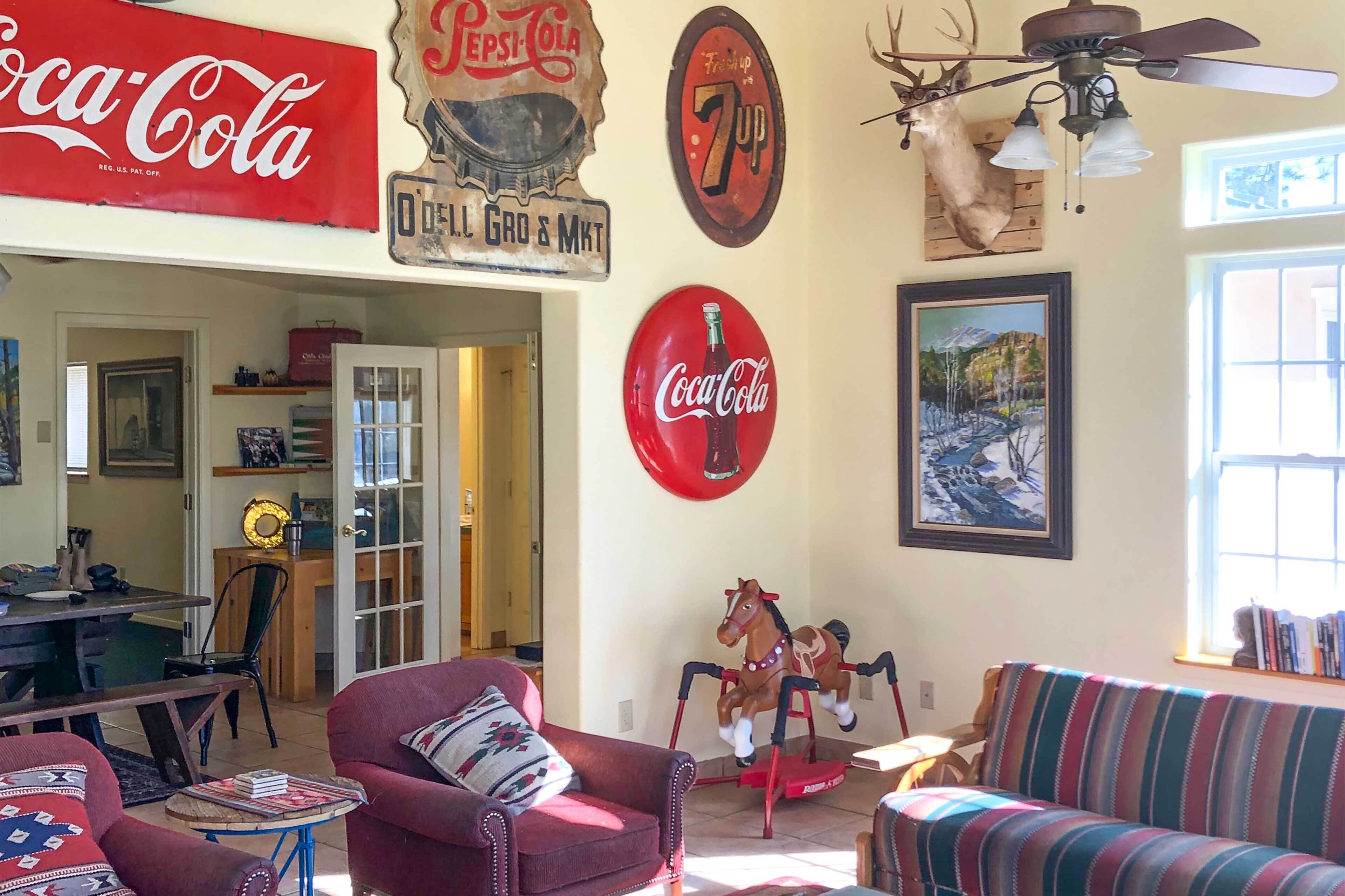 Una sala de estar con un letrero rojo de Coca-Cola, un letrero vintage de 7-Up, una cabeza de venado y un caballito mecedor de Coca-Cola.