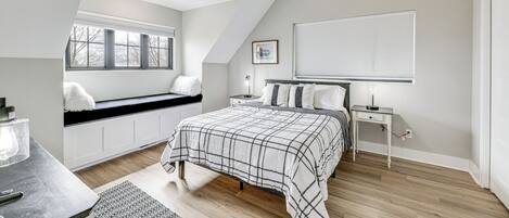 A bedroom with a sloped ceiling, a bed with a plaid bedspread, a window seat, a bedside table with a lamp, and a nightstand.