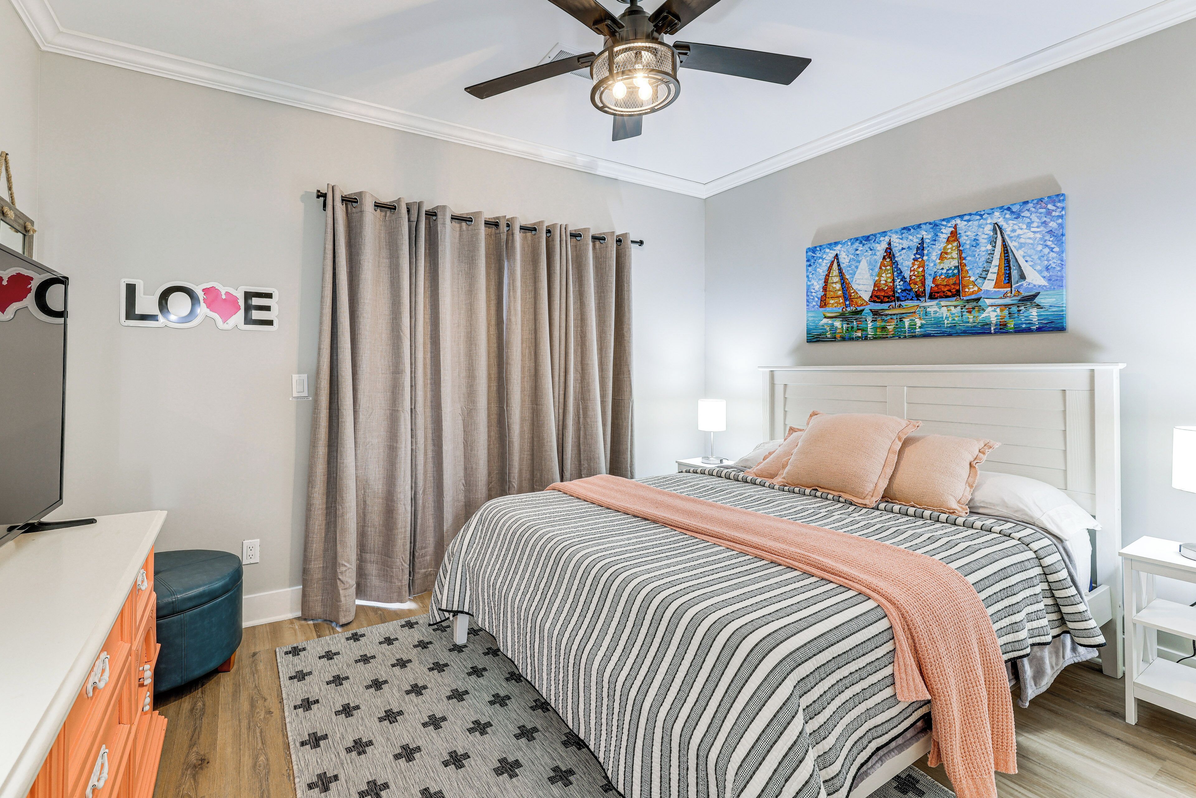 Une chambre à coucher avec un lit, des rideaux, un ventilateur de plafond, un tableau et une commode.