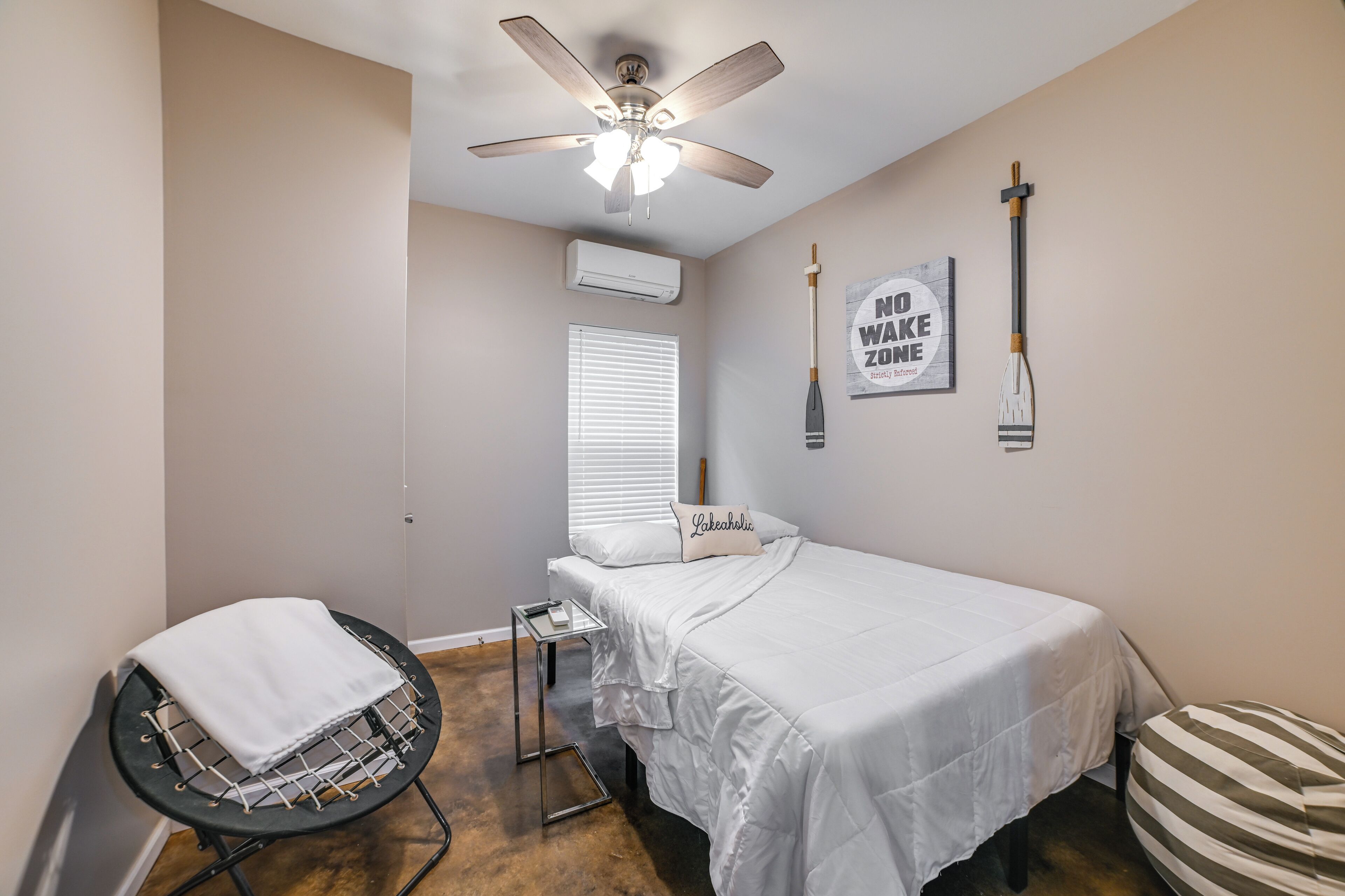 A bedroom with a bed, a ceiling fan, a window with blinds, a wall-mounted air conditioner, and a motivational poster.