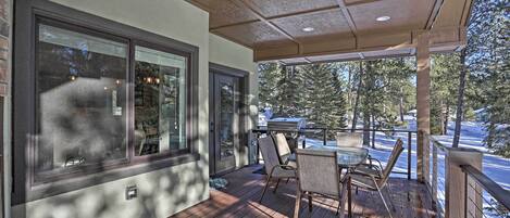 A covered outdoor deck with a table and chairs, large windows, and a view of trees and a car.