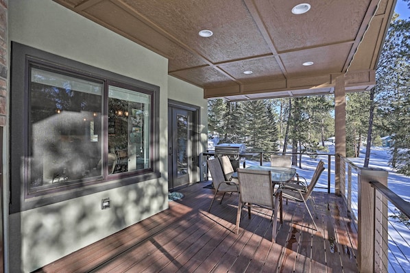 A covered outdoor deck with a dining table and chairs, large windows, and a view of trees and a vehicle.