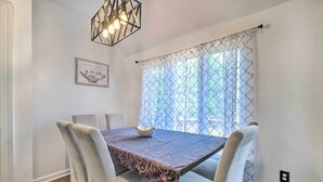 A dining area with a table set for four, a decorative light fixture above, and a window with patterned curtains. - Pond-view Deck + Pool Table: Woodbridge Haven! (Woodbridge)