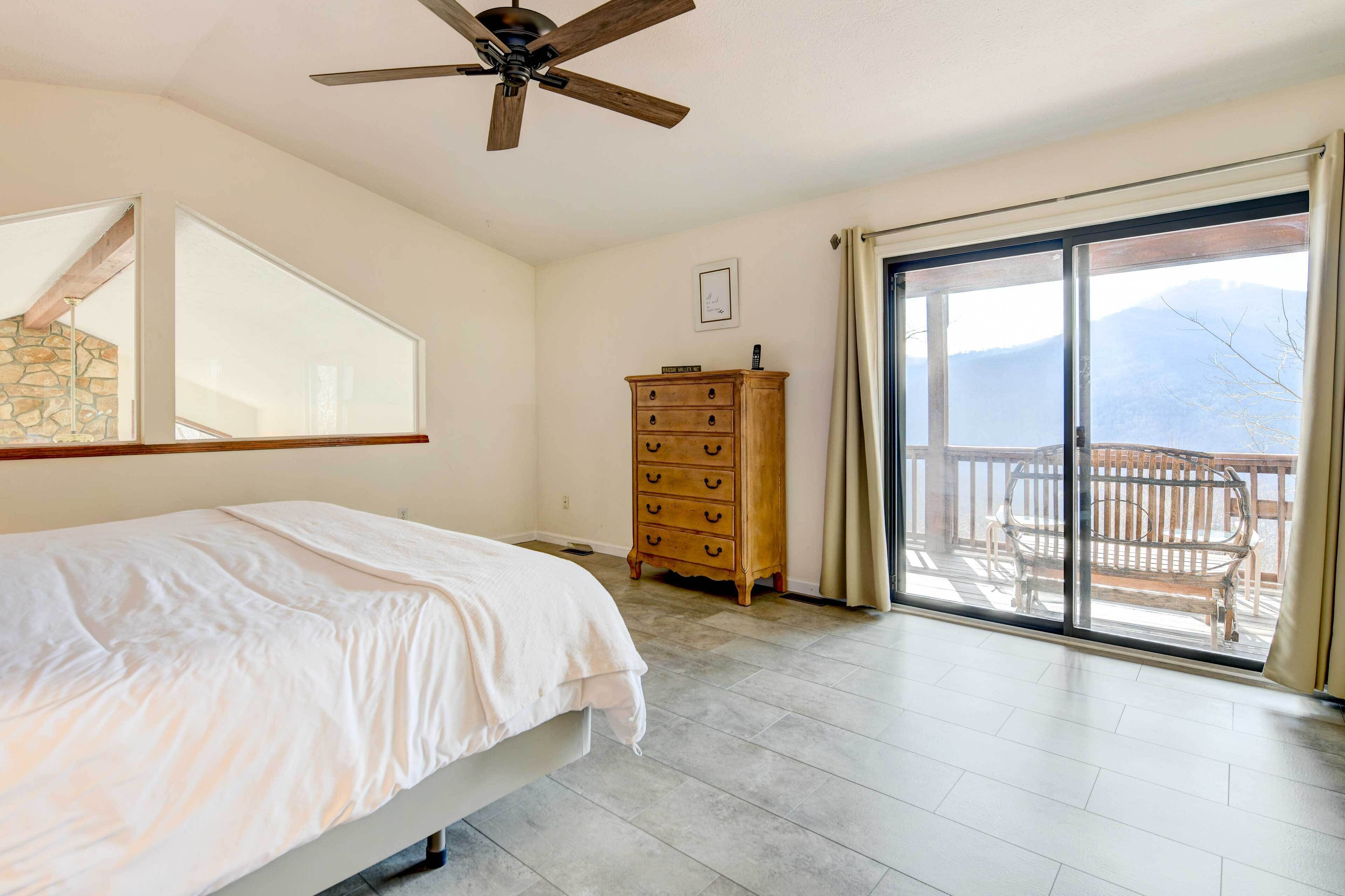 A bedroom with a bed, a wooden dresser, a ceiling fan, and a sliding glass door leading to a balcony with a view of mountains.