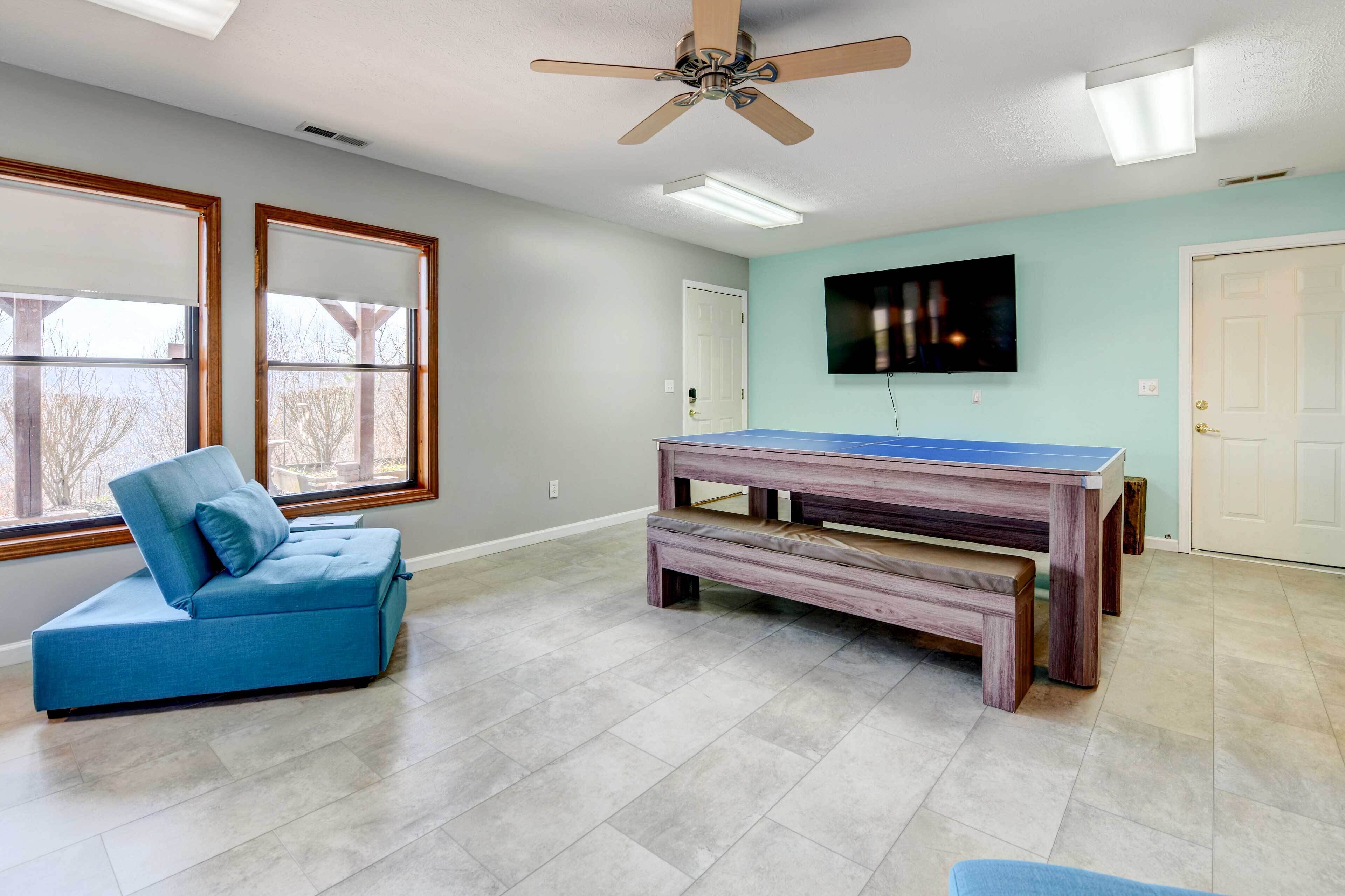 A room with a blue sofa, a ping pong table, a ceiling fan, and a TV on the wall.