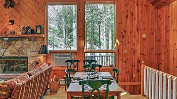 A cozy cabin interior with a stone fireplace, wooden walls, a dining area with a table and chairs, and a large window overlooking a forest.