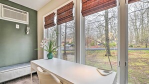 Una habitación con un escritorio blanco, una silla, una planta en maceta y ventanas grandes con persianas marrones.