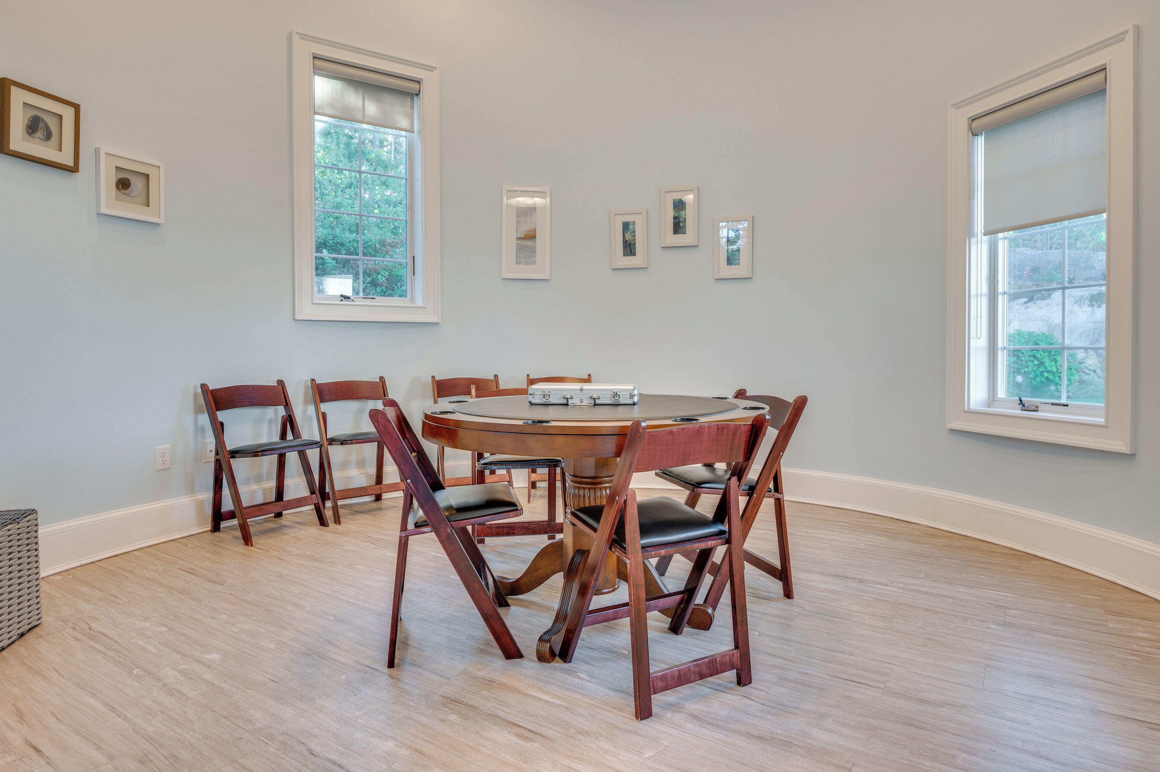 A dining room with a round wooden table and six chairs, large windows, and framed pictures on the wall.