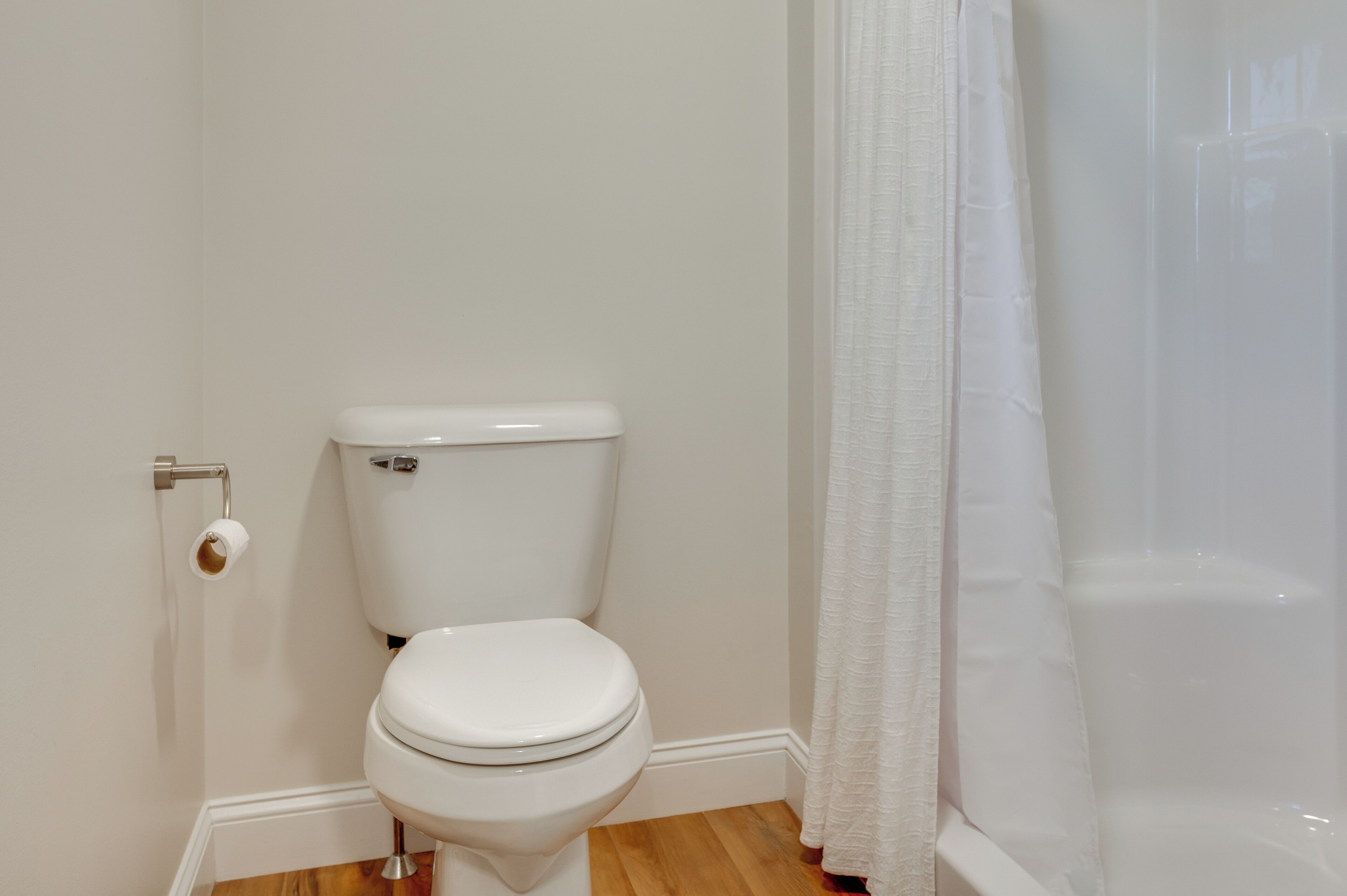 Des toilettes blanches avec un porte-serviettes en papier, un rideau de douche blanc et un plancher en bois.