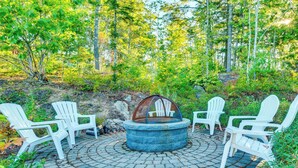 A circular stone fire pit with a glass enclosure, surrounded by four white Adirondack chairs in a forested area with a paved stone floor. - Stylish Arundel Cottage, 6 Mi to Dock Square! (Kennebunkport)