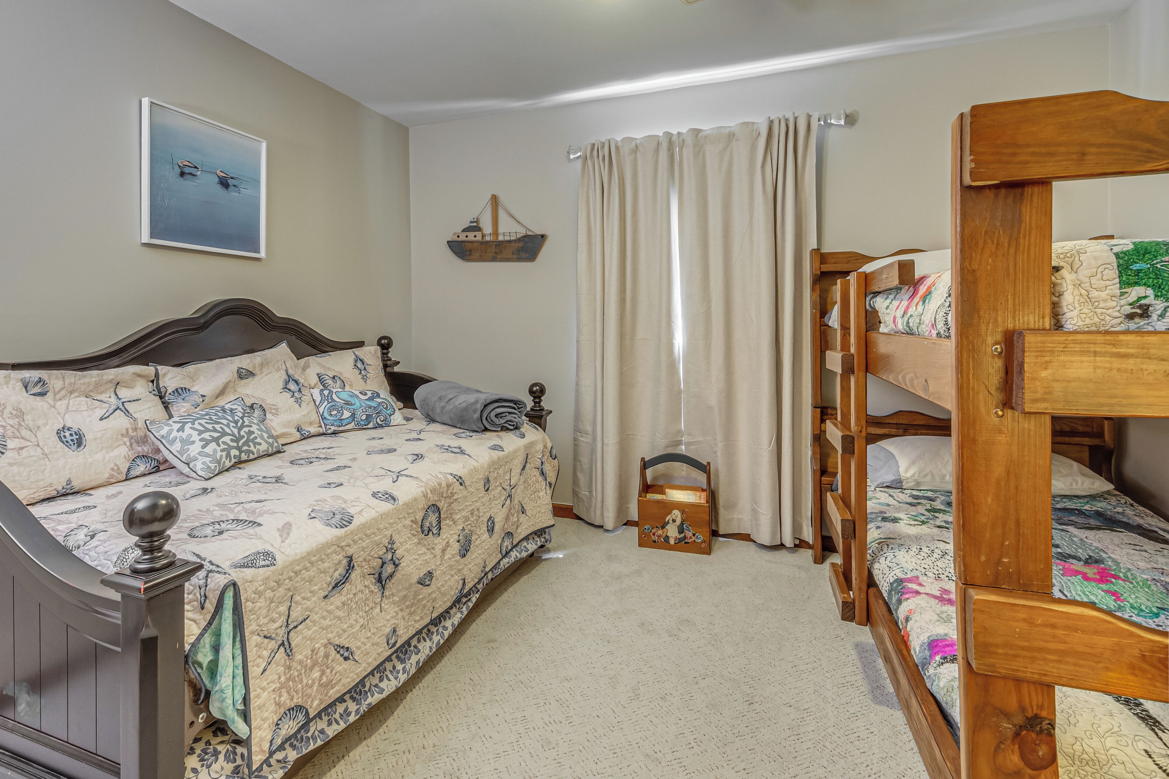 Un dormitorio con cama, cómoda, mesita de noche, litera de madera, un adorno de barco colgante y una ventana con cortinas.