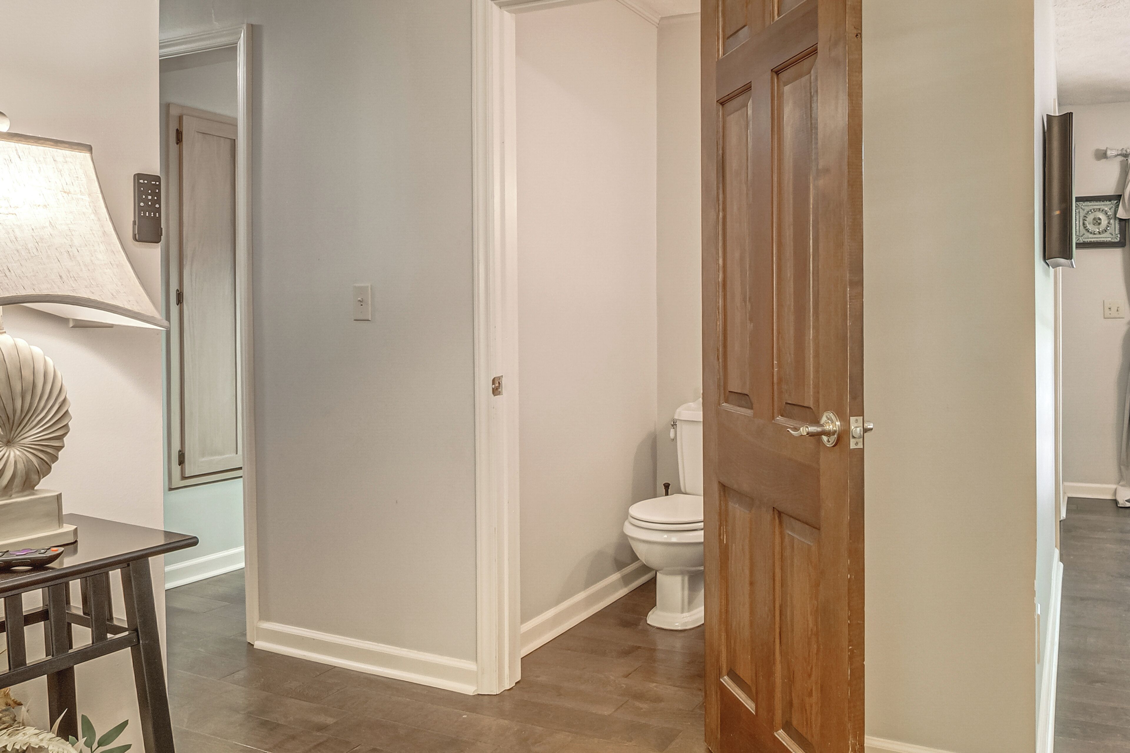 Un couloir avec une porte en bois, des toilettes blanches et une lampe posée sur une table.