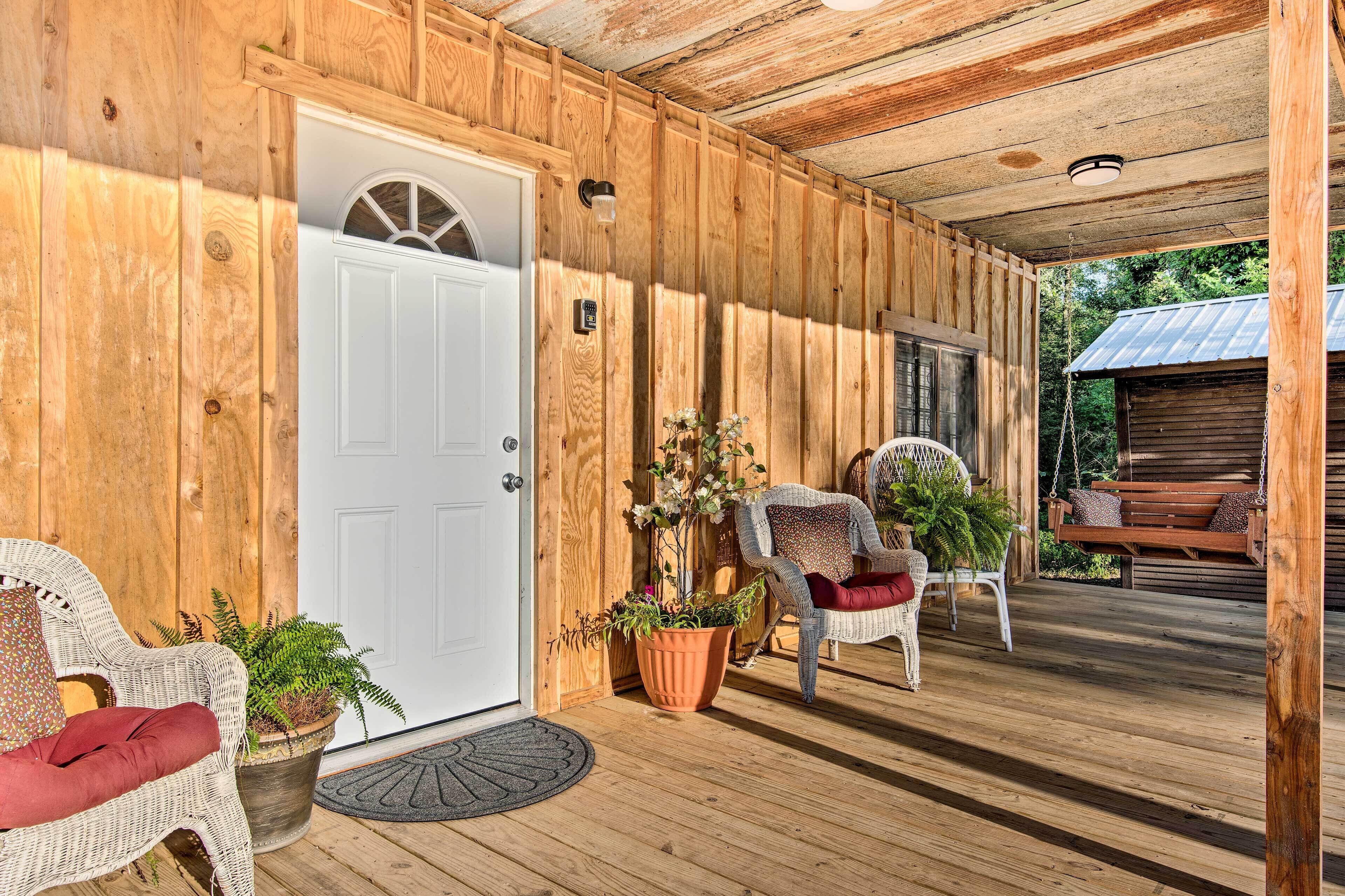 A wooden porch with a white door, wicker chairs with cushions, and potted plants.