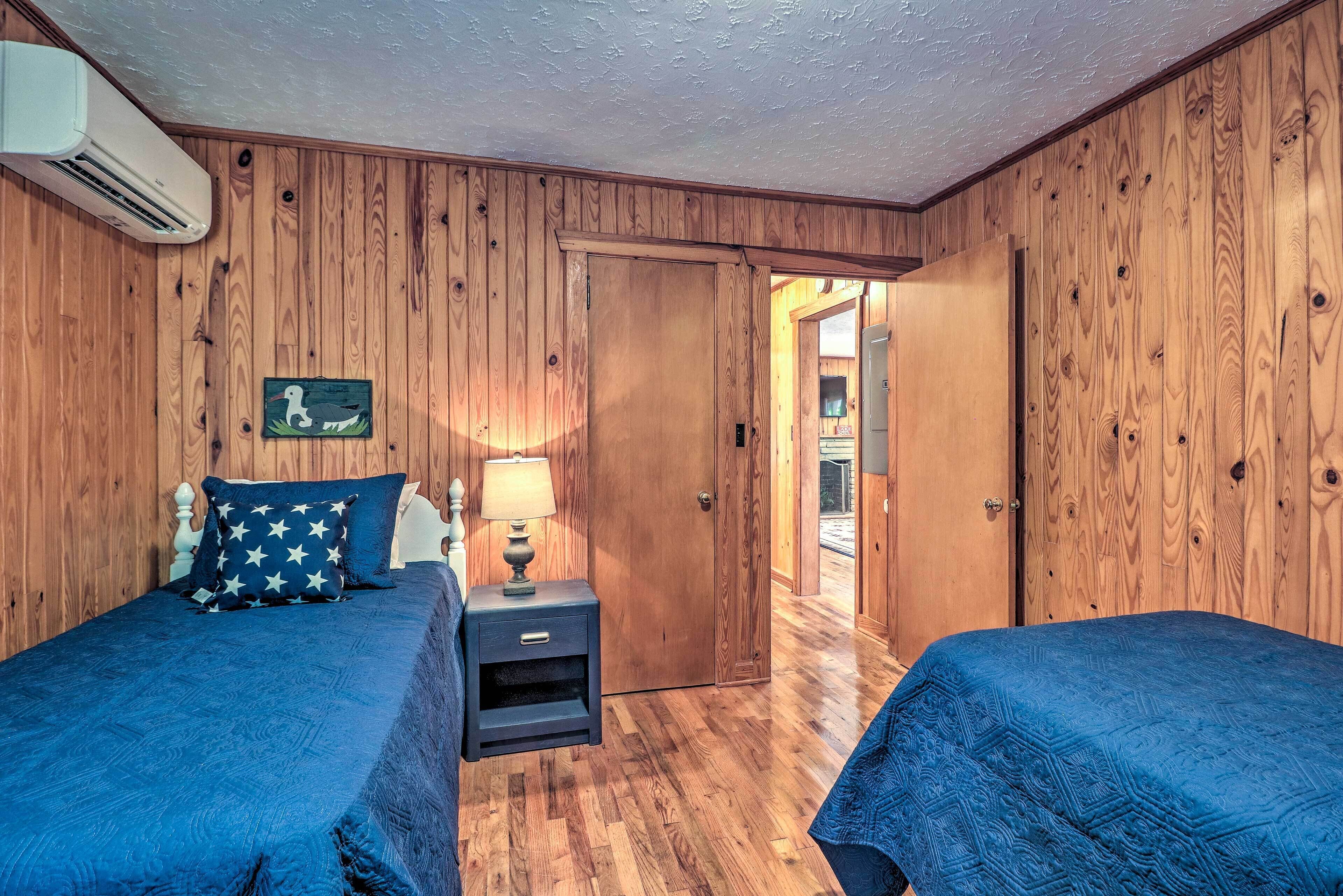 Una habitación con paredes y puerta de madera. Hay dos camas con ropa de cama azul y una mesita de noche con una lámpara.