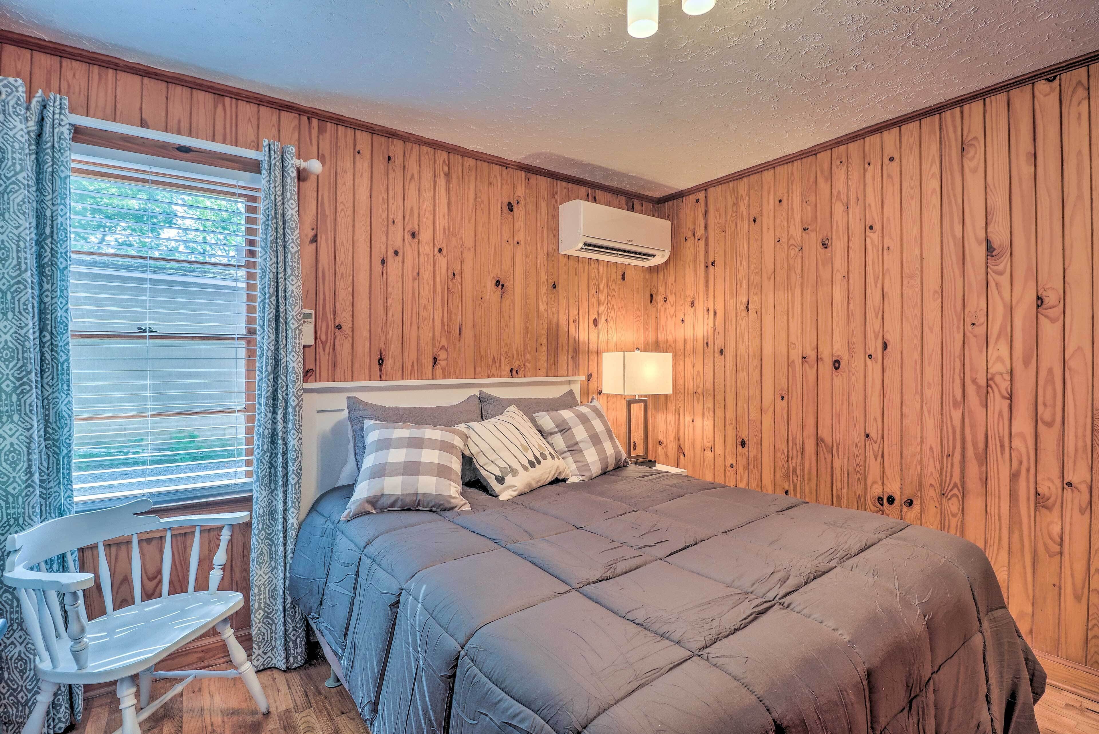 A bedroom with a wooden paneled wall, a bed with a grey duvet, a white chair, and a window with blinds.