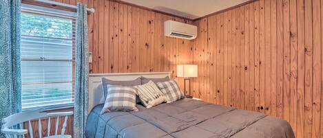 A bedroom with a wooden paneled wall, a bed with a grey duvet, a white chair, and a window with blinds.
