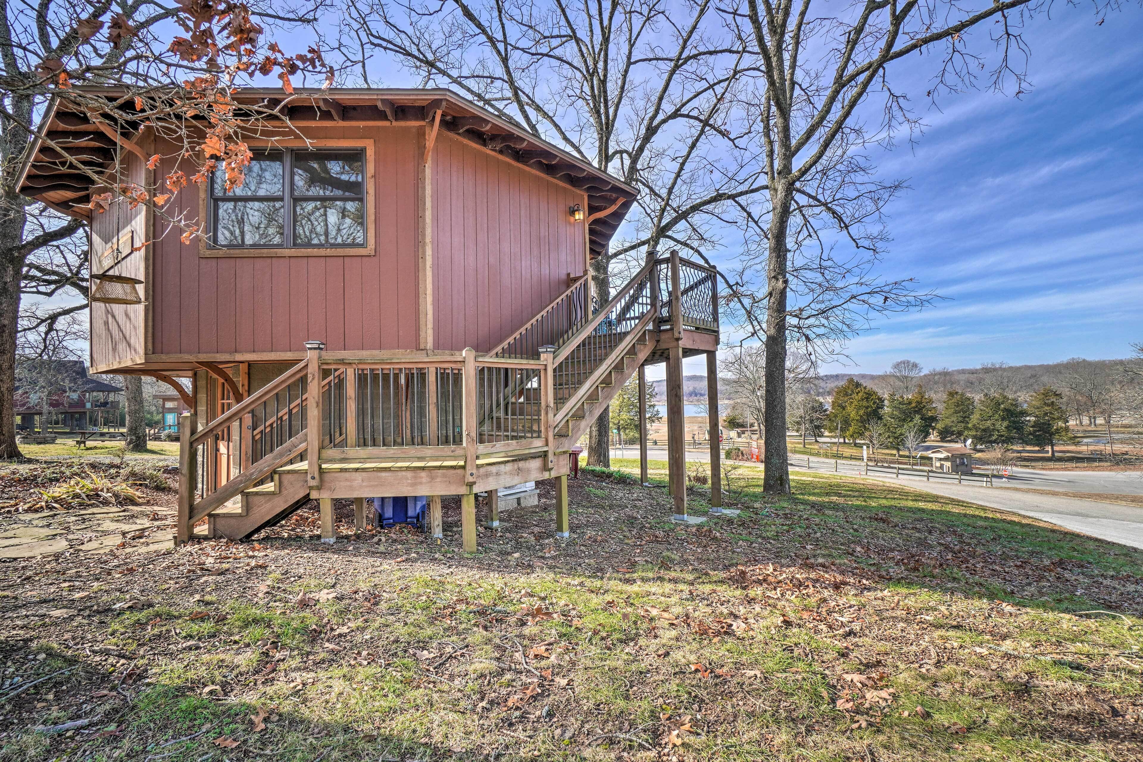 A wooden cabin with a deck and stairs, surrounded by trees and a clear sky.