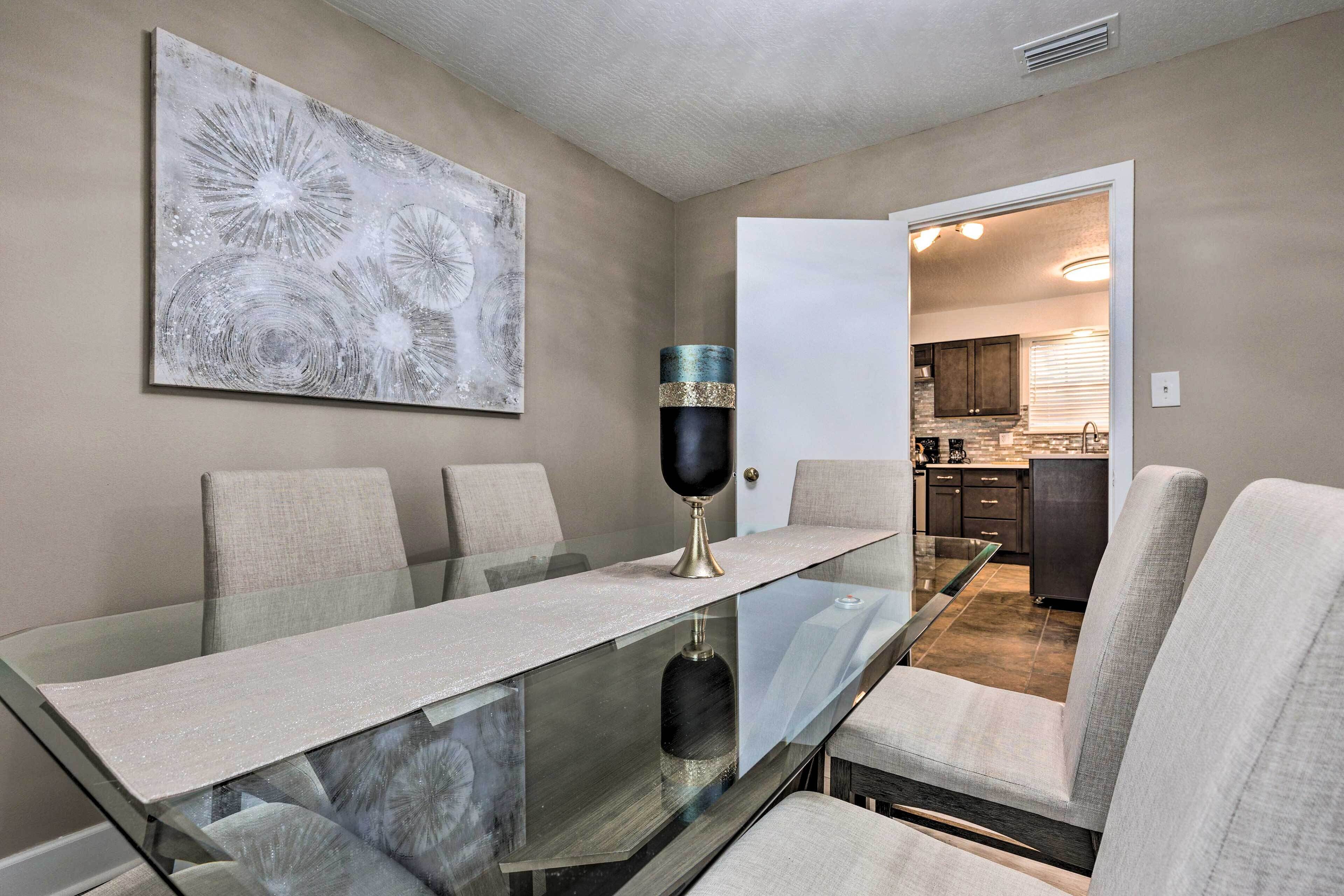 Un comedor con una mesa de vidrio, cuatro sillas, un jarrón decorativo y un cuadro abstracto grande en la pared.