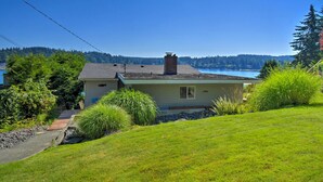 A single-story house with a chimney, a small deck, and a view of a lake and forested hills.