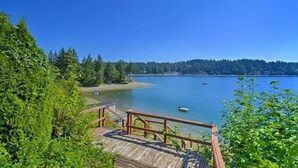 A wooden deck with a railing overlooking a calm lake with boats, surrounded by dense forest and a clear blue sky.