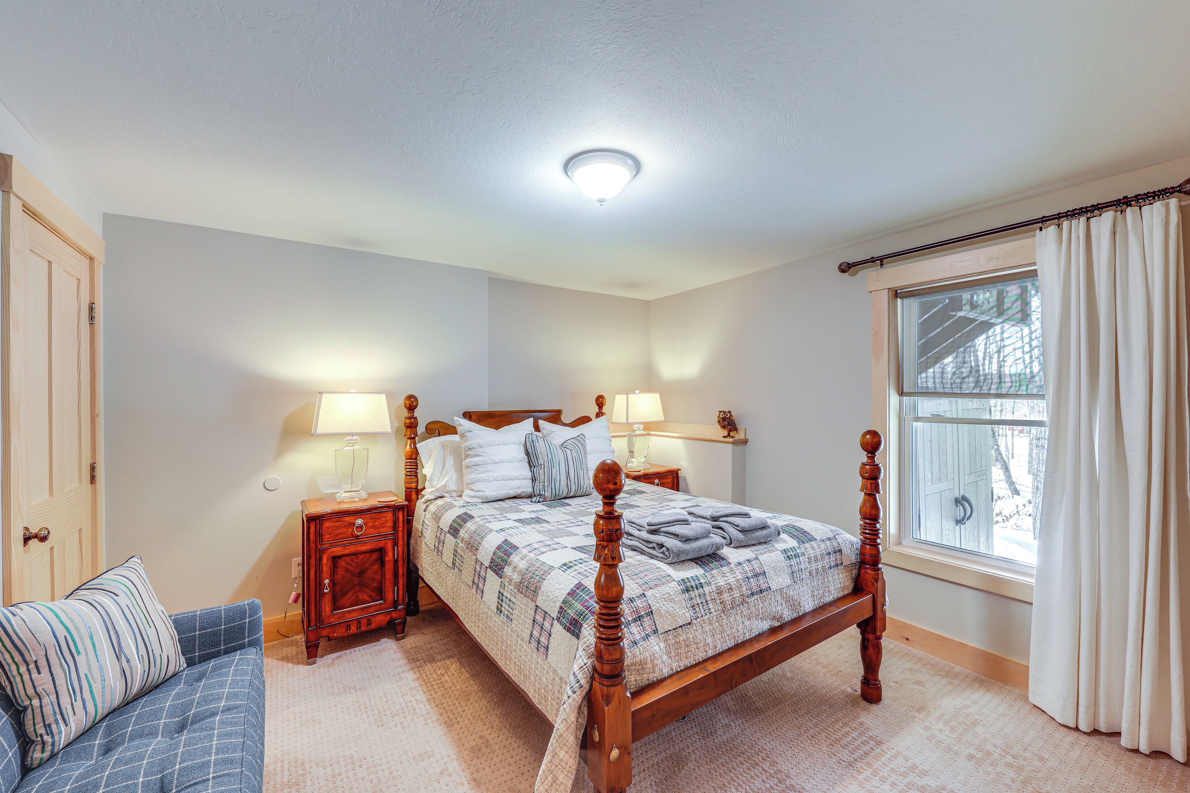 A bedroom with a wooden four-poster bed, two bedside tables with lamps, a blue sofa, and a window with curtains.