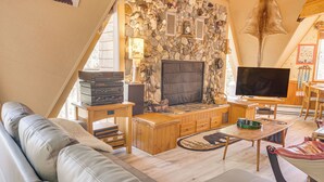 A cozy living room with a stone wall, a fireplace, a television, and a leather sofa. - 'star House' Unique Home w/ 360 Mountain Views! (Pine Mountain Club)
